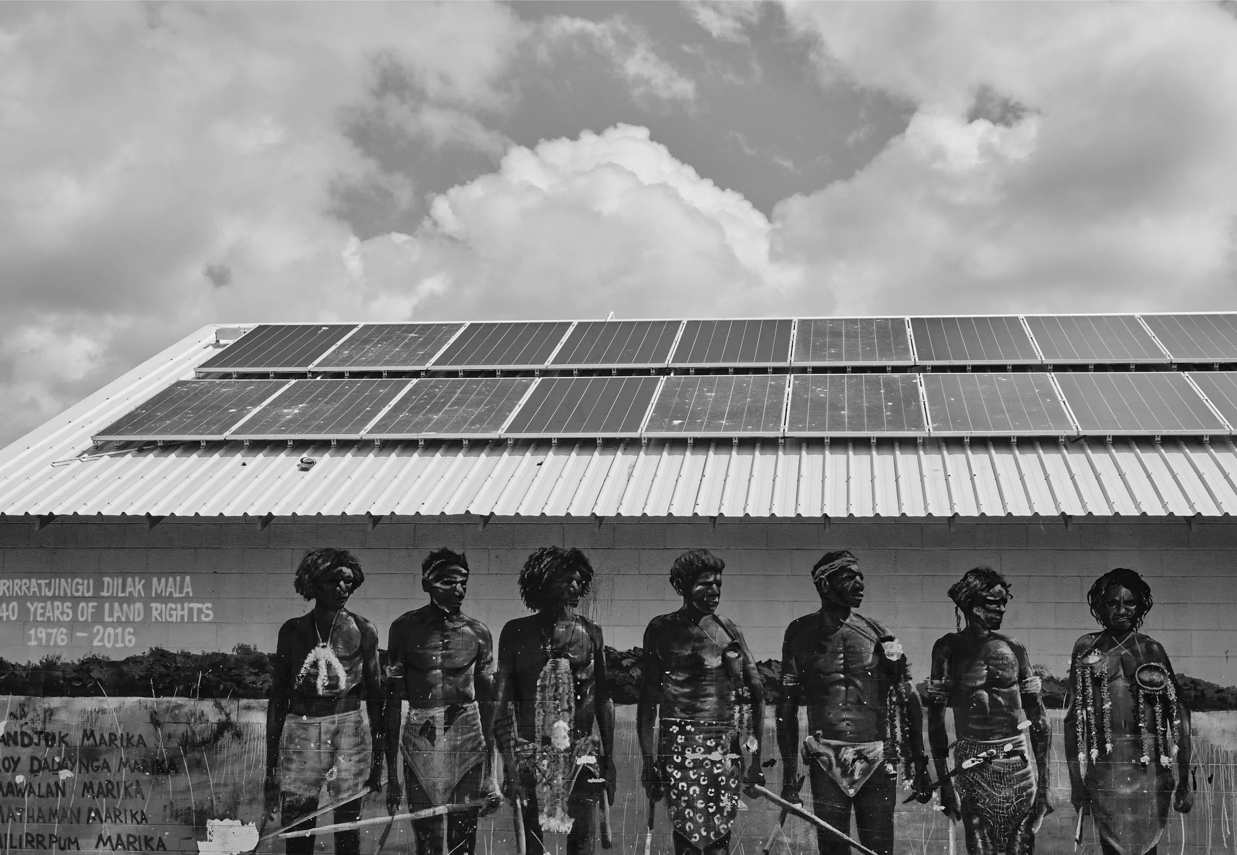 A group of indigenous women standing in a line, holding sticks, with a building that has solar panels on the roof and a mural in the background.