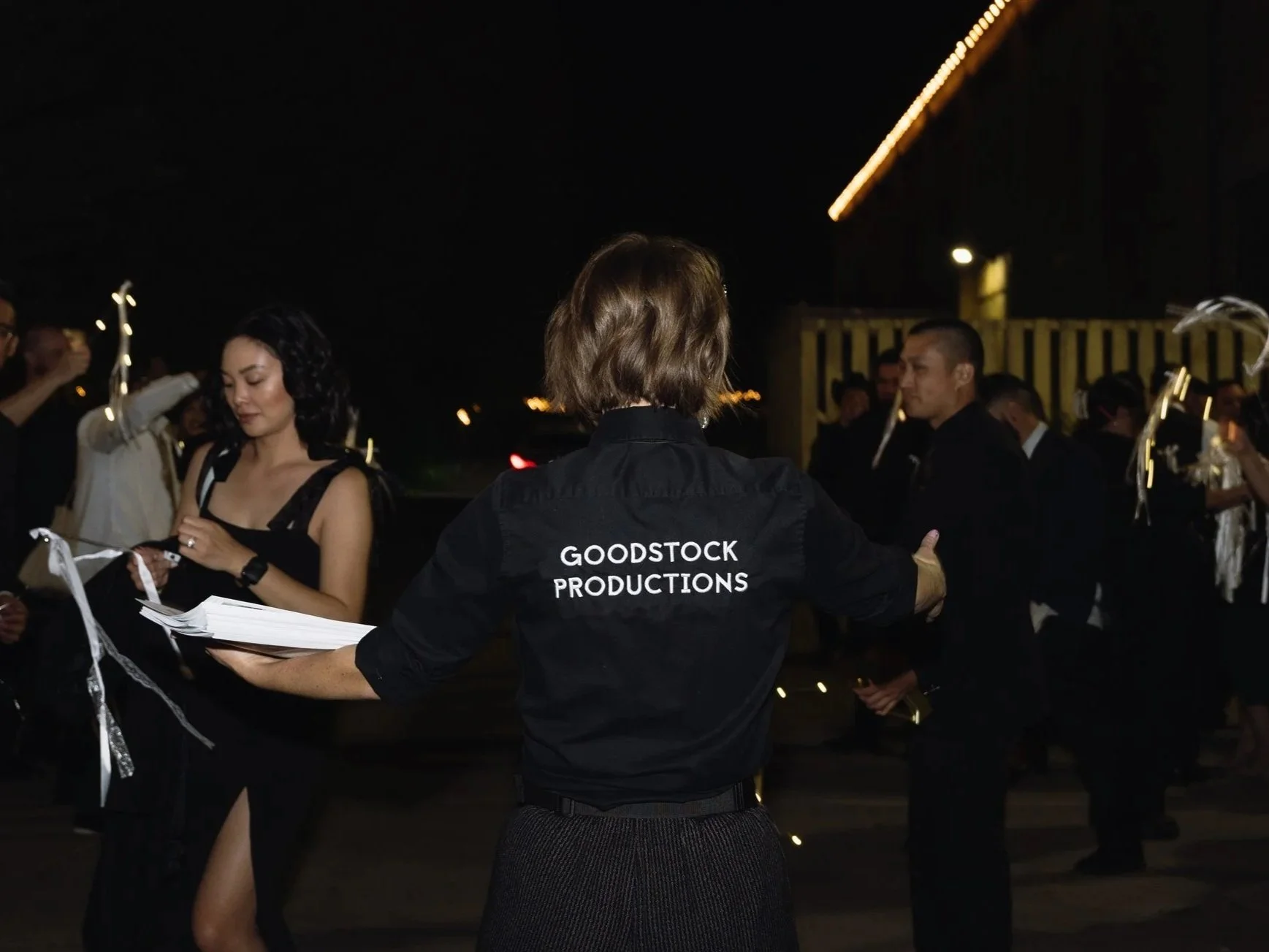 Night scene of people at an event, with a woman in a black dress holding papers and another person with 'Goodstock Productions' on their jacket in the foreground, and other attendees with white feather centerpieces in the background.