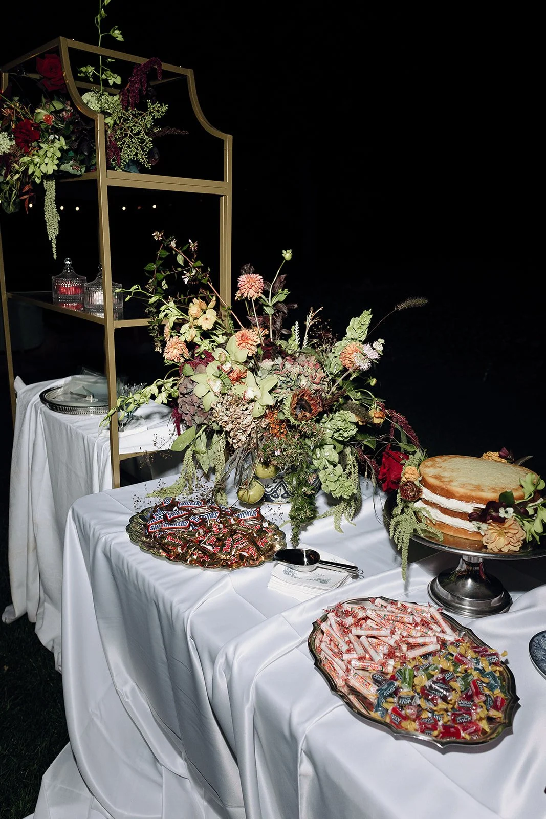 A dessert table with a large wedding cake, bowls of candy, and floral arrangement with draping