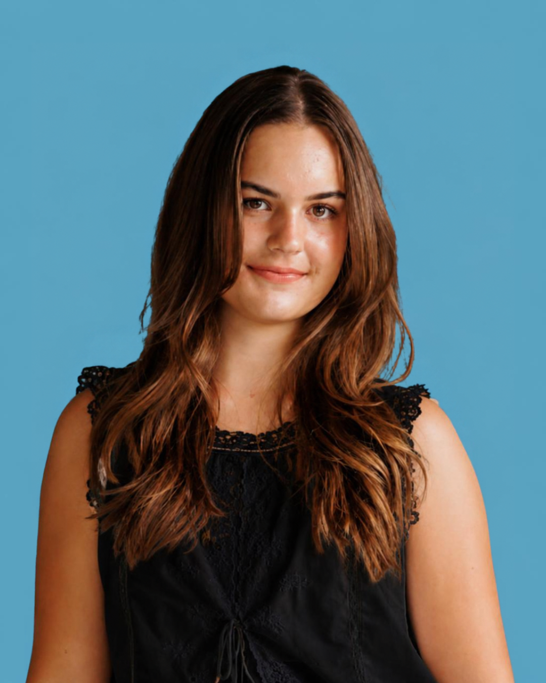 A young woman with long, wavy brown hair wearing a black sleeveless top standing against a light blue background.
