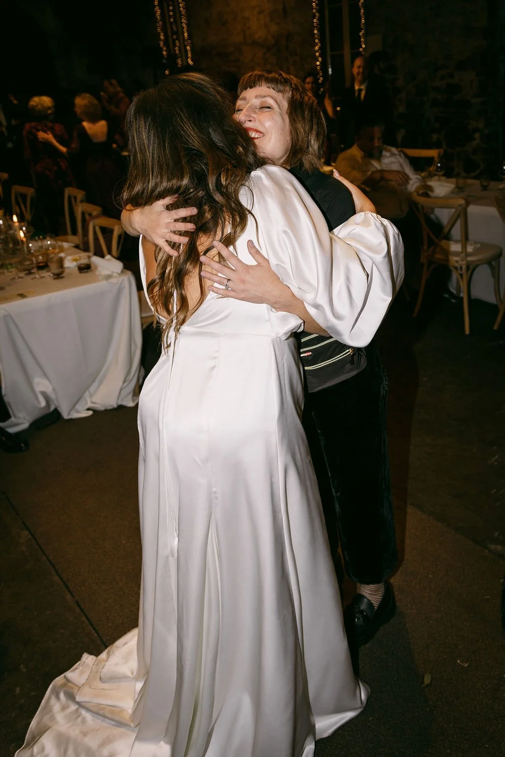 Bride hugging her wedding planner in a warmly lit event space with tables and guests in the background.