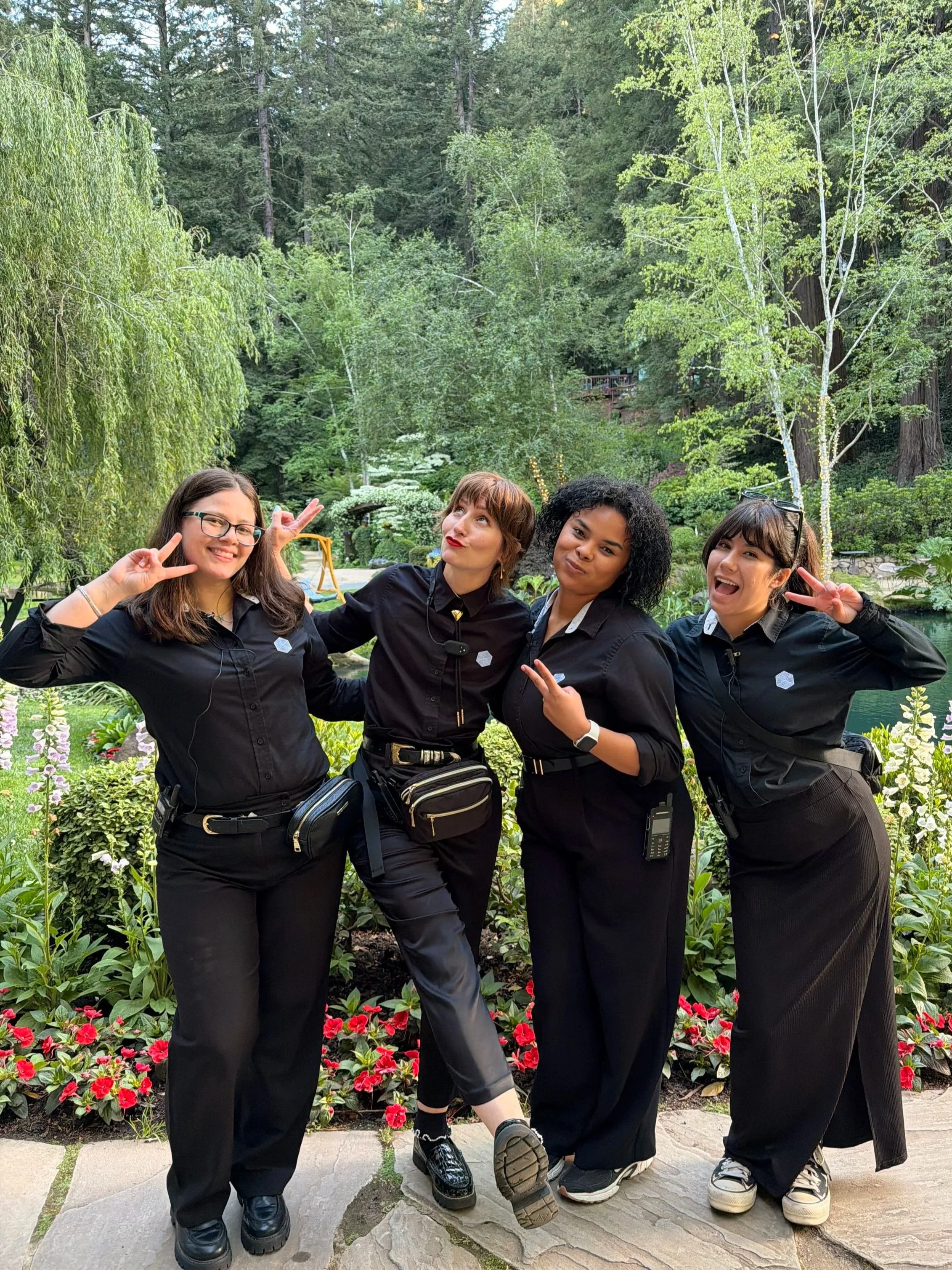 Wedding planning team of four women posing with peace signs and smiles in a lush, green garden with trees and red flowers.