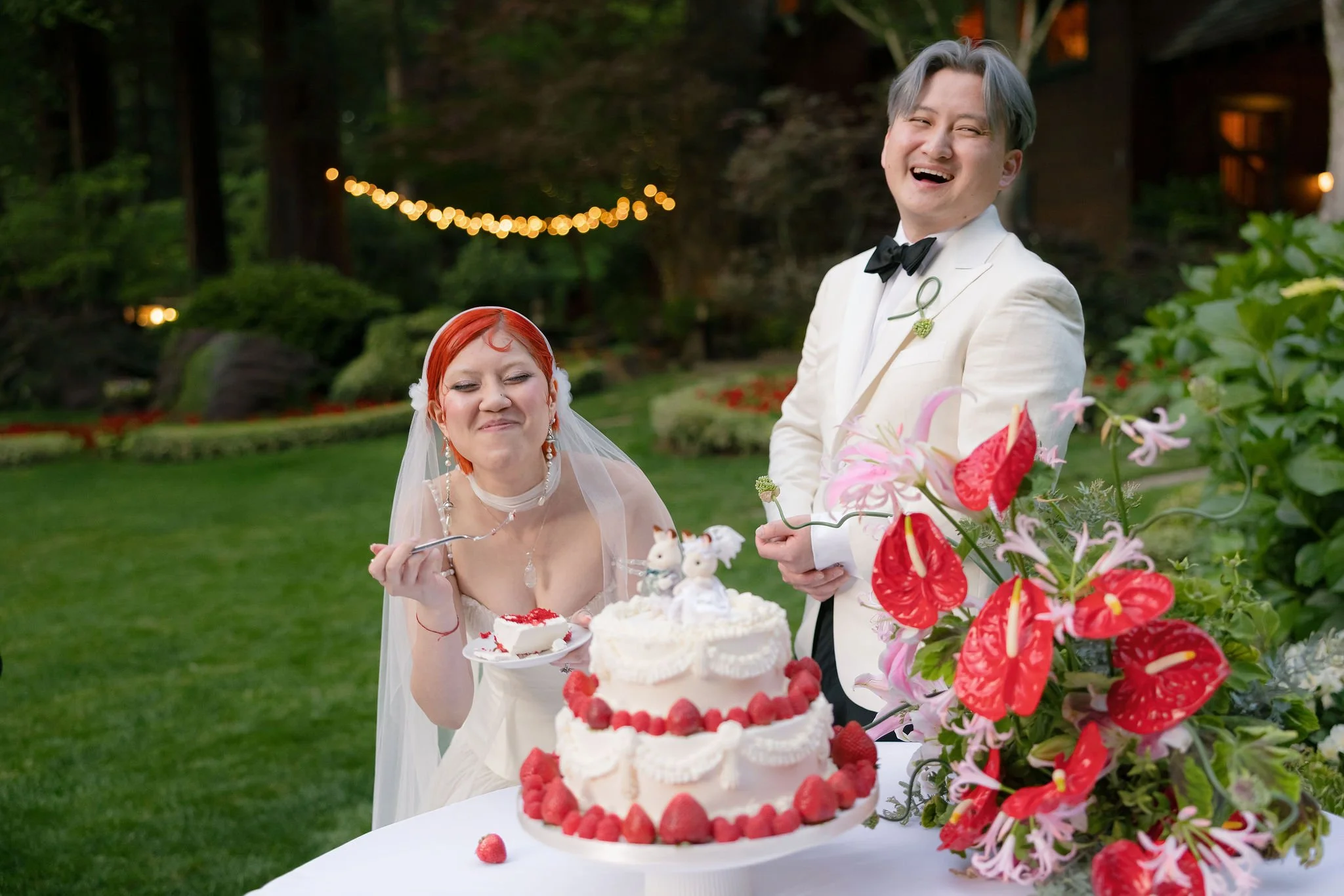 A joyful wedding celebration outdoors featuring a bride and groom in formal attire, with the bride in a white dress and the groom in a cream tuxedo with a black bow tie. They are smiling and preparing to cut awedding cake decorated with strawberries, with a table full of flowers in the foreground