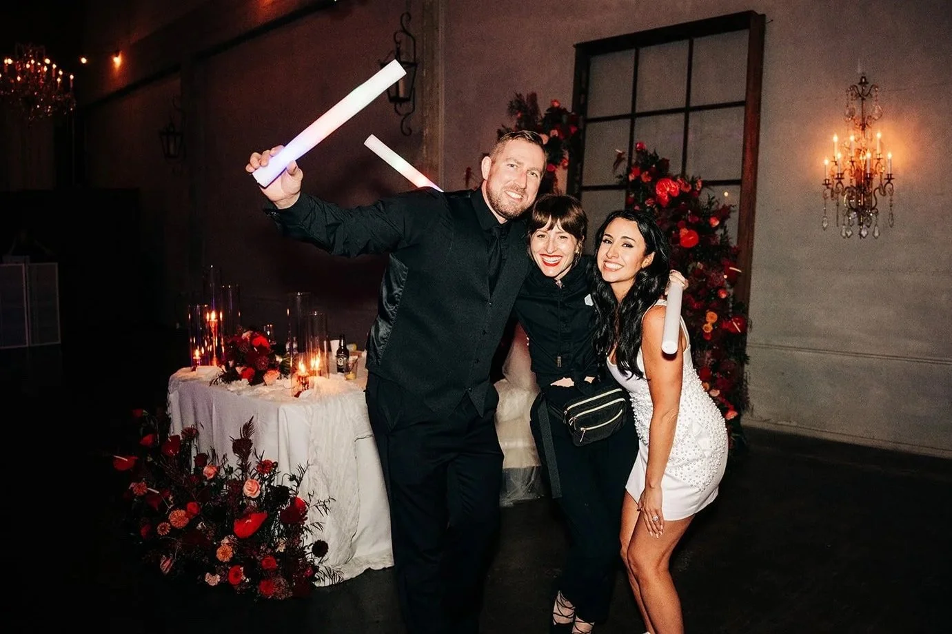 Three people celebrating at a party with LED foam sticks, a decorated table with candles, flowers, and drinks, and a background with a flower arrangement and wall sconces.