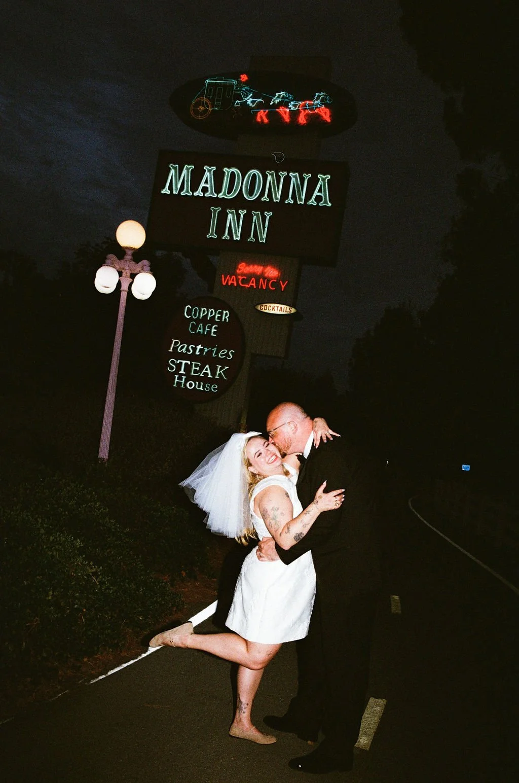 A couple in wedding attire dancing outside at night in front of the Madonna Inn sign, which is illuminated with neon lights and displays the inn's name and amenities such as Copper Cafe, Pastries, Steak House, and a sign indicating vacancies.