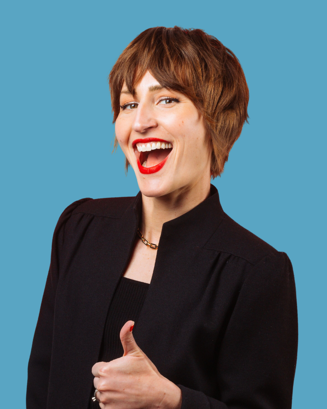 A woman with short red hair wearing a black blazer and red lipstick, smiling and giving a thumbs-up gesture against a blue background.