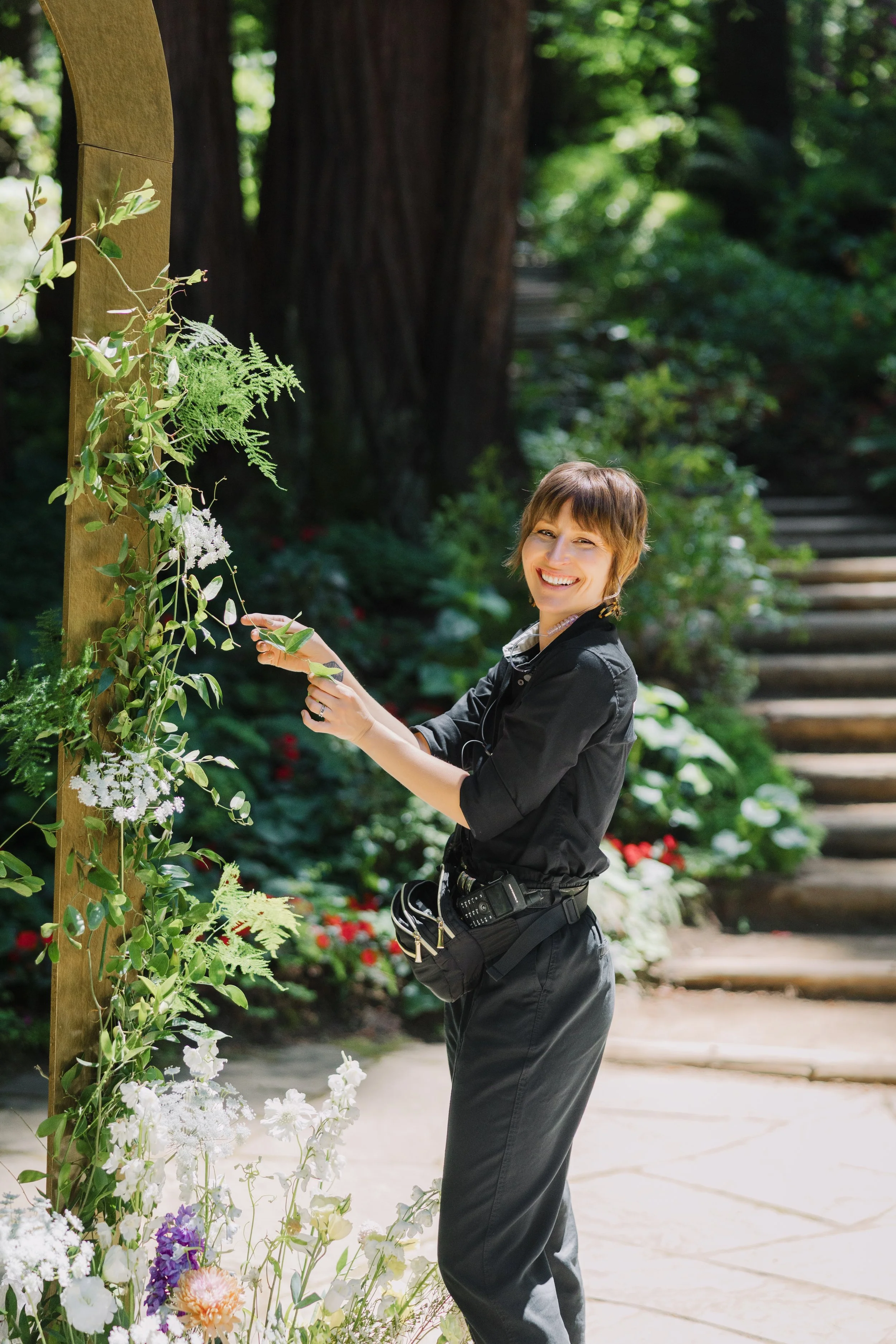 A wedding planner smiling and holding a flower stem in a garden, standing next to a wooden arch decorated with various flowers. She is in a sunny outdoor setting with trees and a pathway nearby.