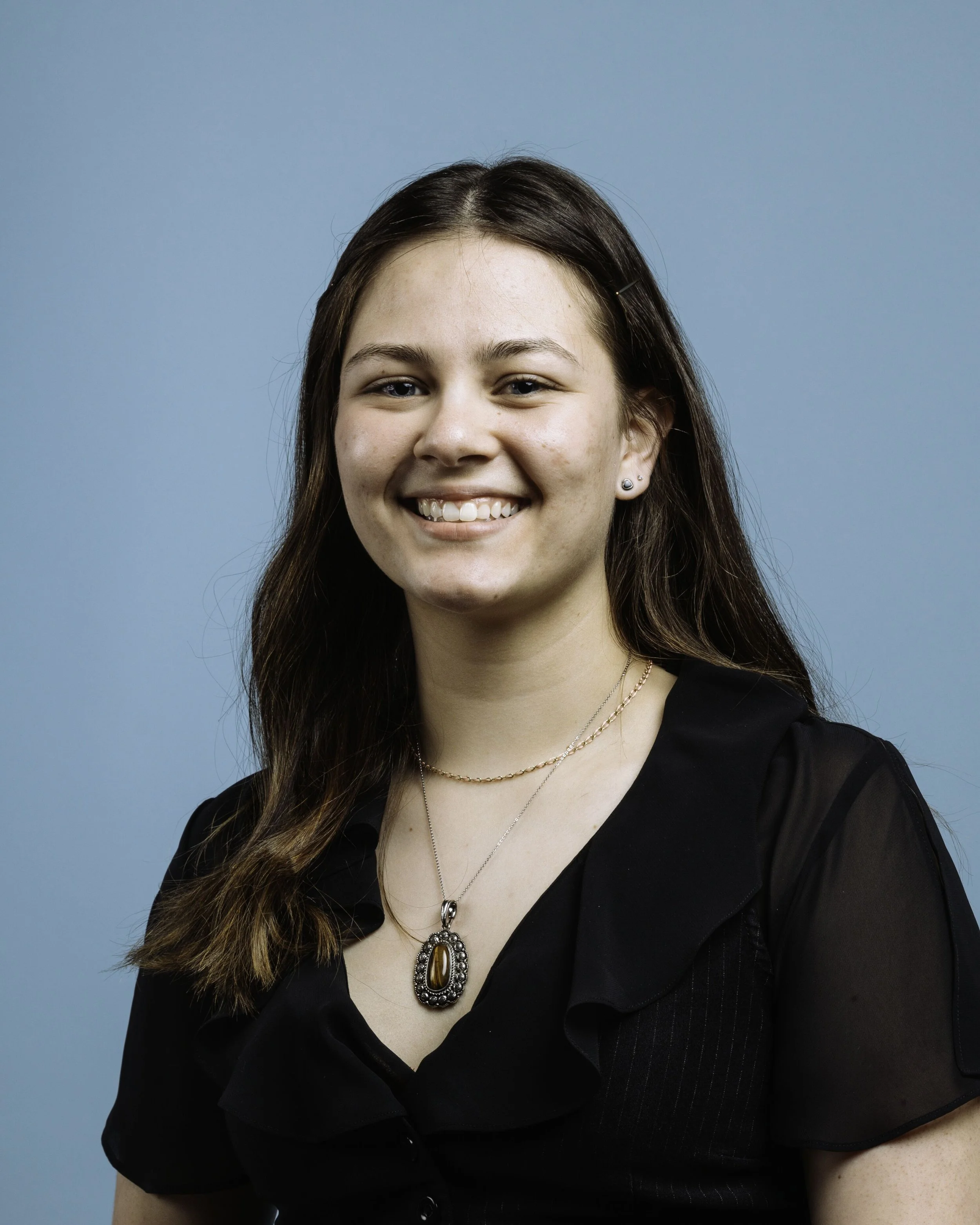 Close-up of a young woman with long brown hair, wearing a black top with ruffled sleeves, stylish jewelry including a large pendant necklace, earrings, and a thin chain, smiling against a light blue background.