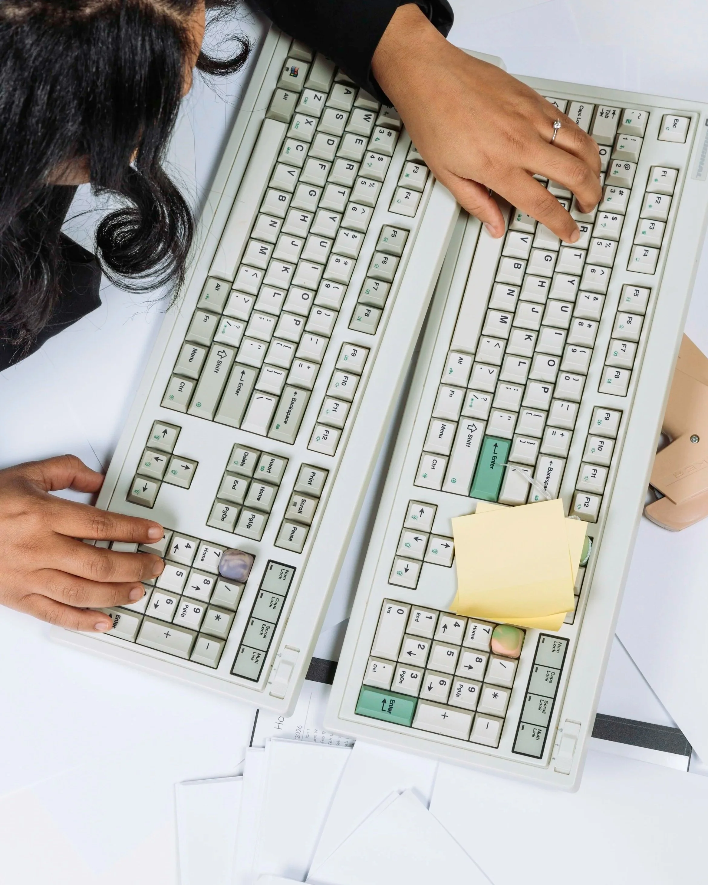 Close-up of two hands typing on vintage computer keyboards with white keys, placed on a desk with scattered papers and sticky notes.