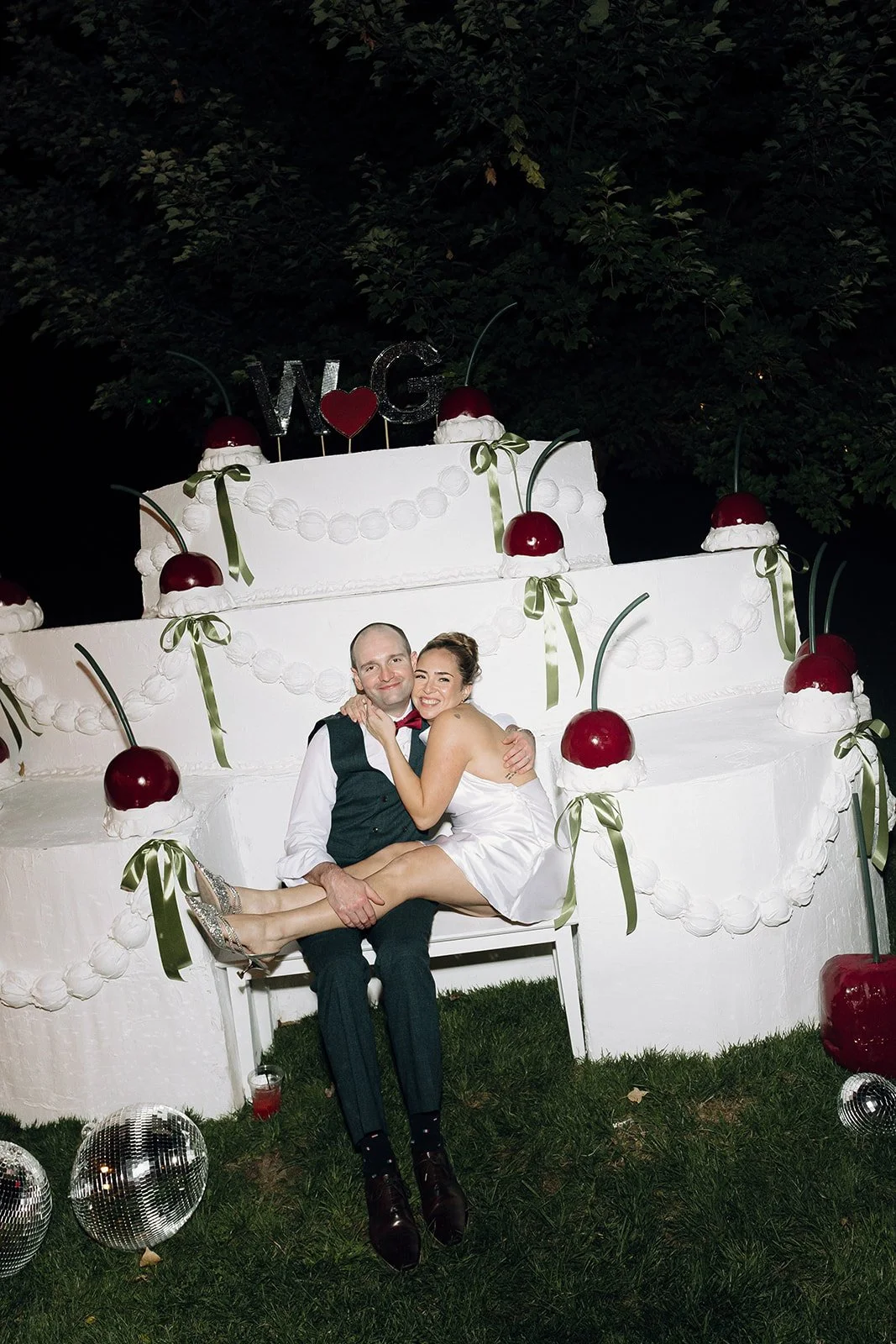 A newlywed couple sitting on a bench in front of a large wedding cake decorated with cherries, green ribbons, and white icing, with a dark outdoor background and disco balls on the grass.