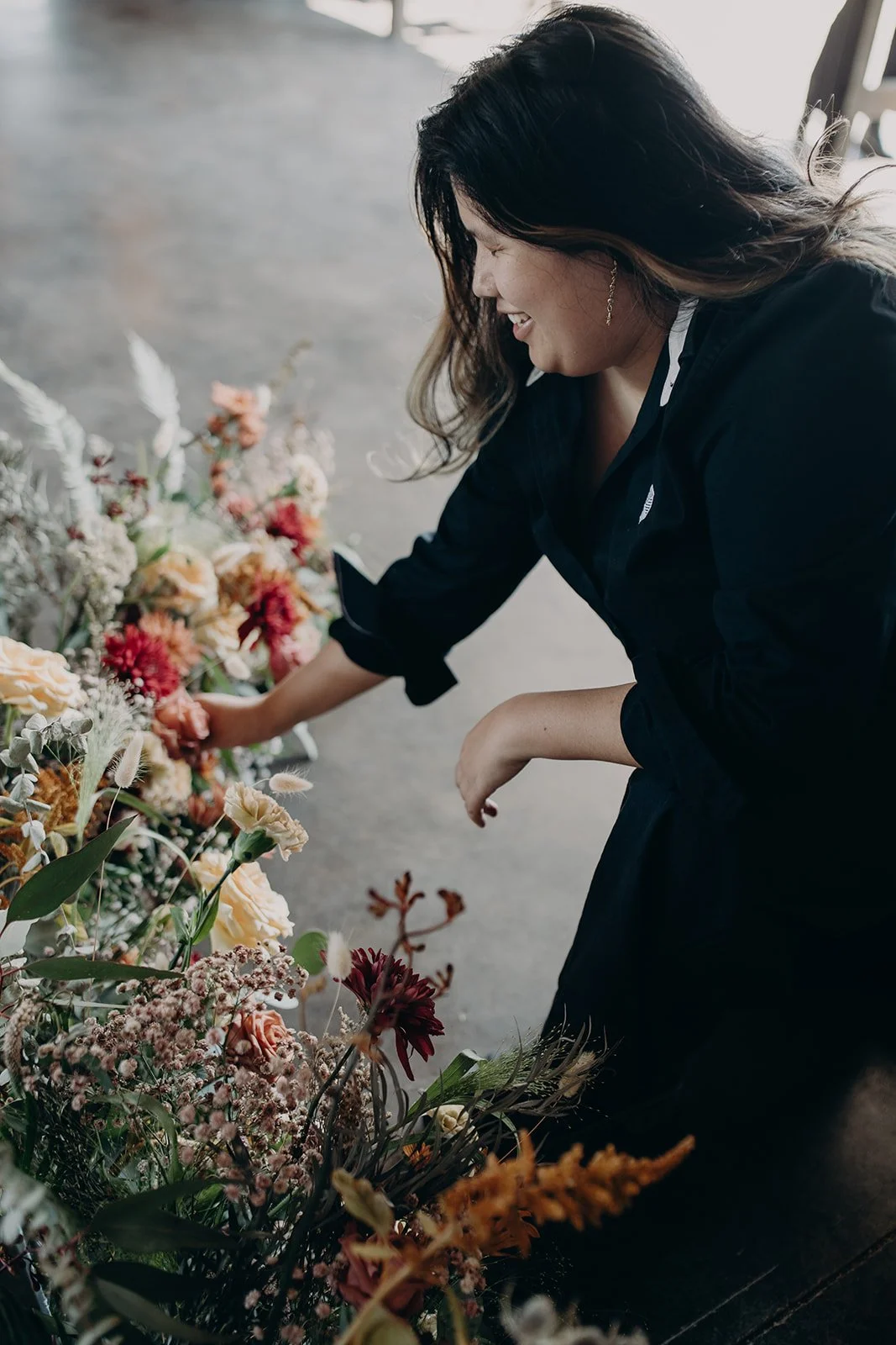 A woman arranging flowers on the ground, smiling and looking at the colorful bouquet.