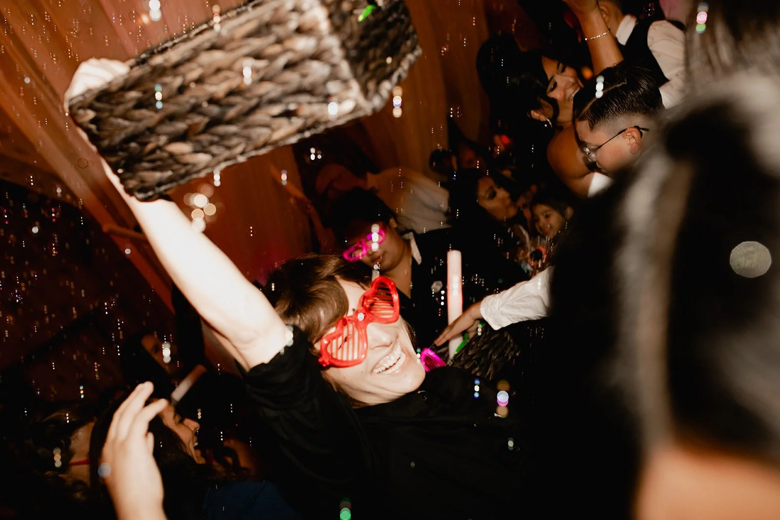 People celebrating at a party with a woman wearing large red heart-shaped glasses in the foreground, surrounded by others dancing and socializing in a decorated room.