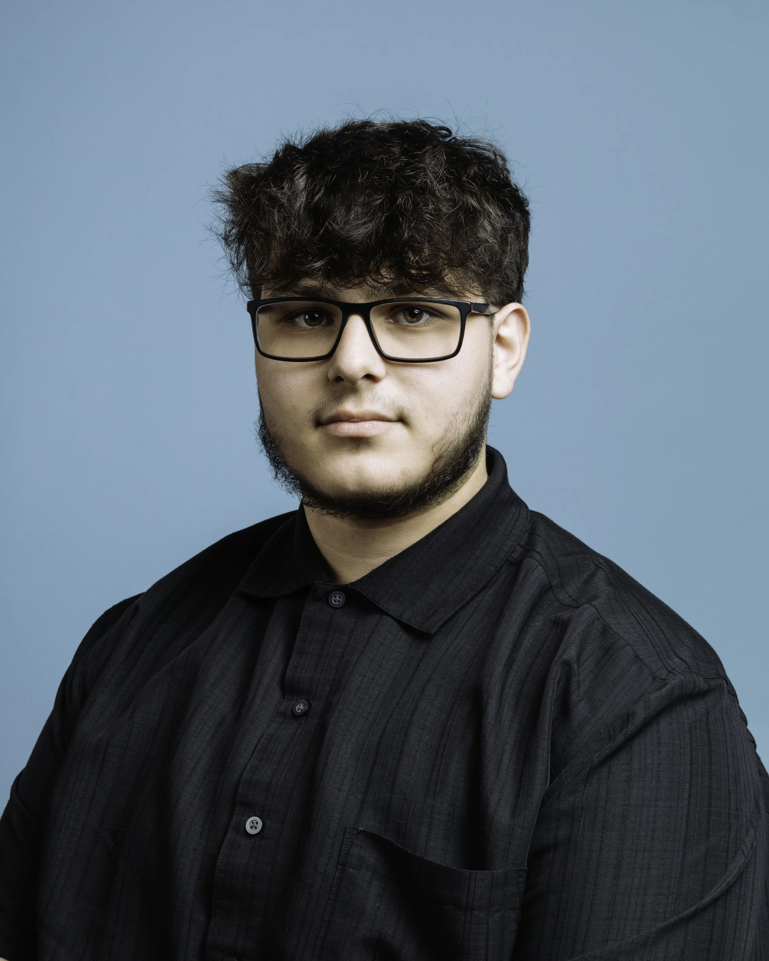 A young man with glasses, short dark hair, and a slight beard wearing a black pinstripe shirt against a blue background.