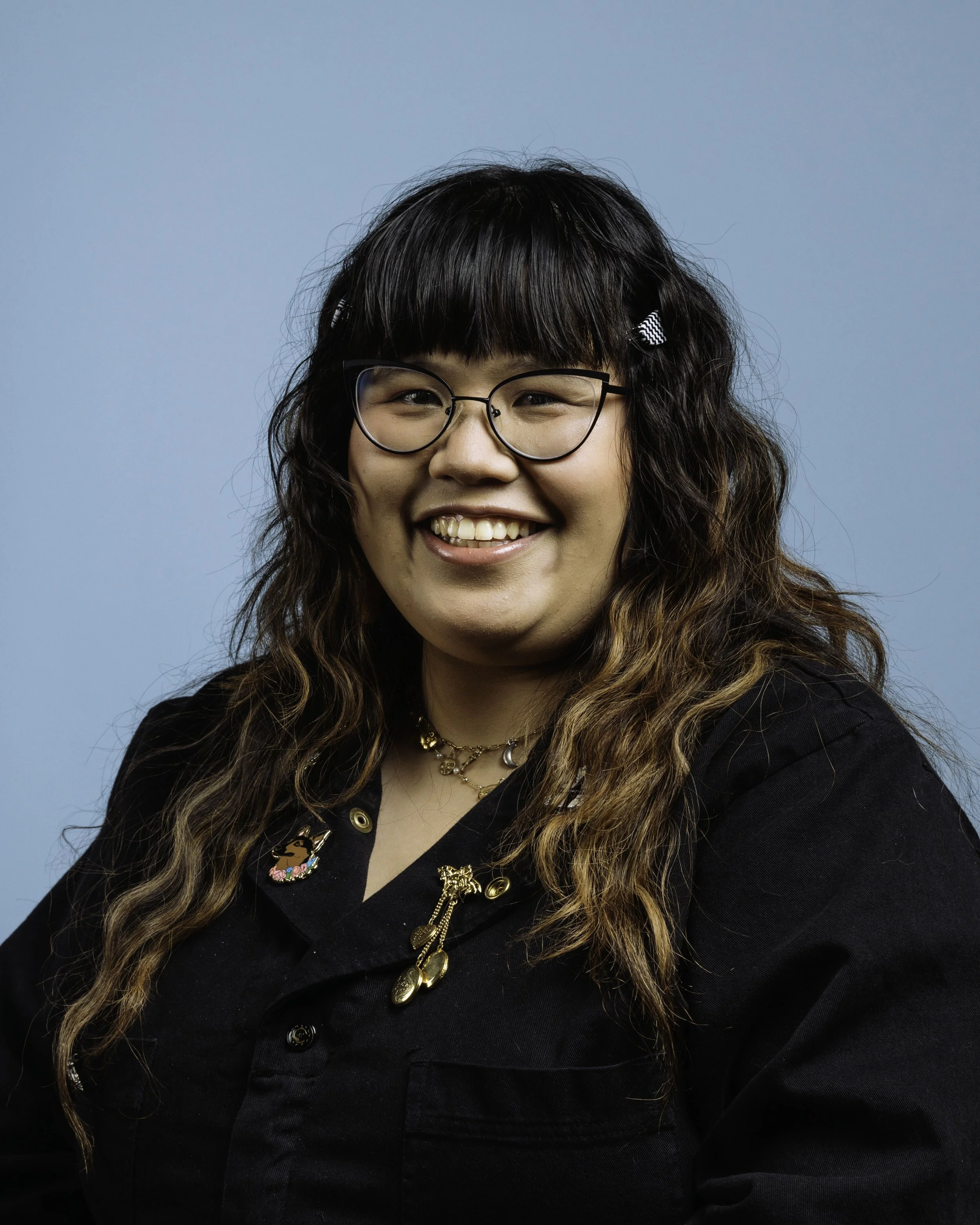 A young woman with long wavy hair, wearing glasses, a black top, and multiple gold jewelry, smiling against a blue background.