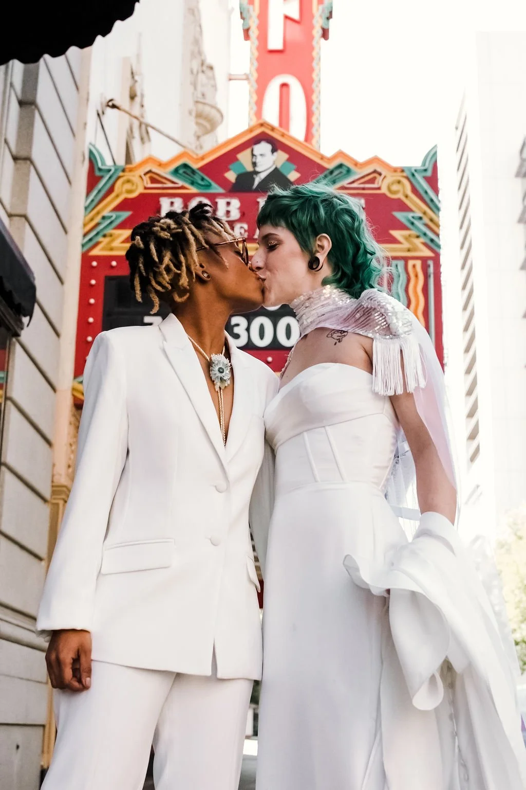 A bride and non-binary partner dressed in white, kissing in front of a neon sign and theater marquee on a city street.
