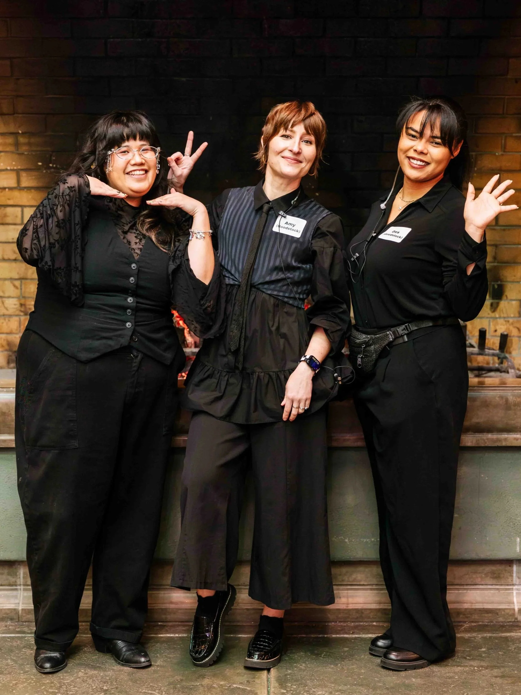 Wedding planning team of three women standing in front of a dark wooden wall, smiling and making playful hand gestures.