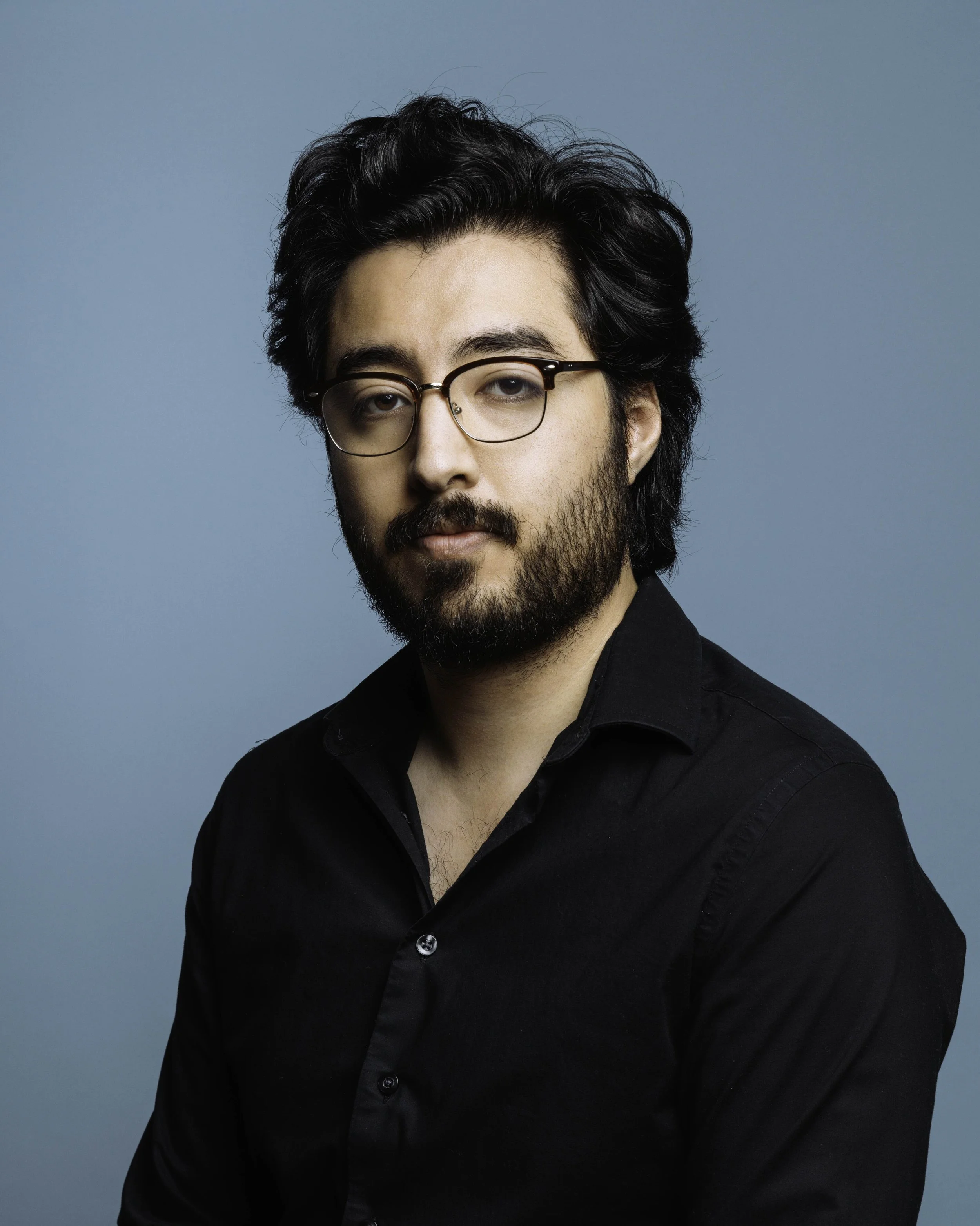 Portrait of a man with dark, wavy hair, glasses, and a beard, wearing a black shirt, against a blue background.