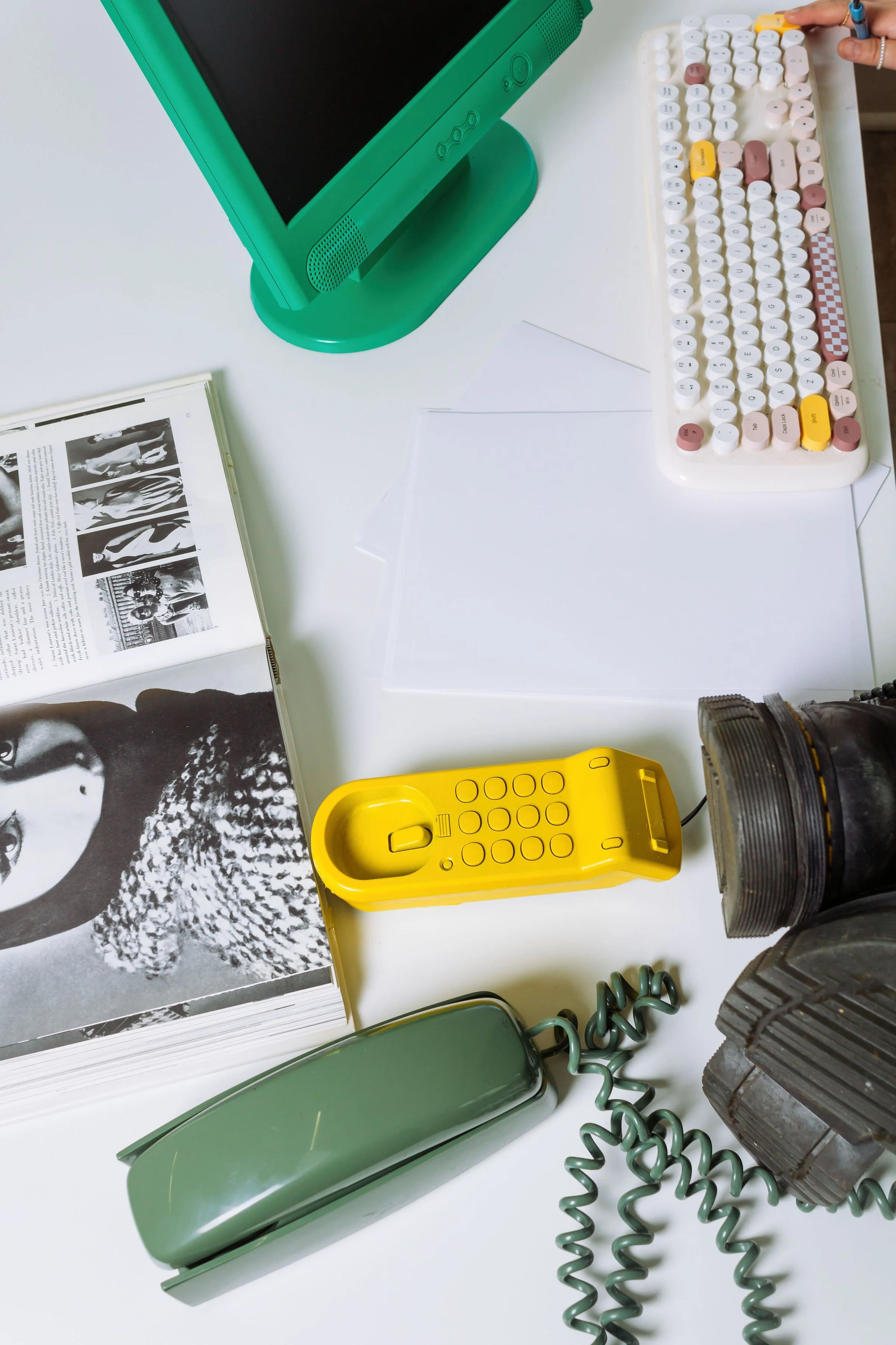 Flat lay of a workspace with a green computer monitor, an open book with black-and-white images, a yellow telephone, a green metal water bottle, a curly green cord, and various objects on a white surface.