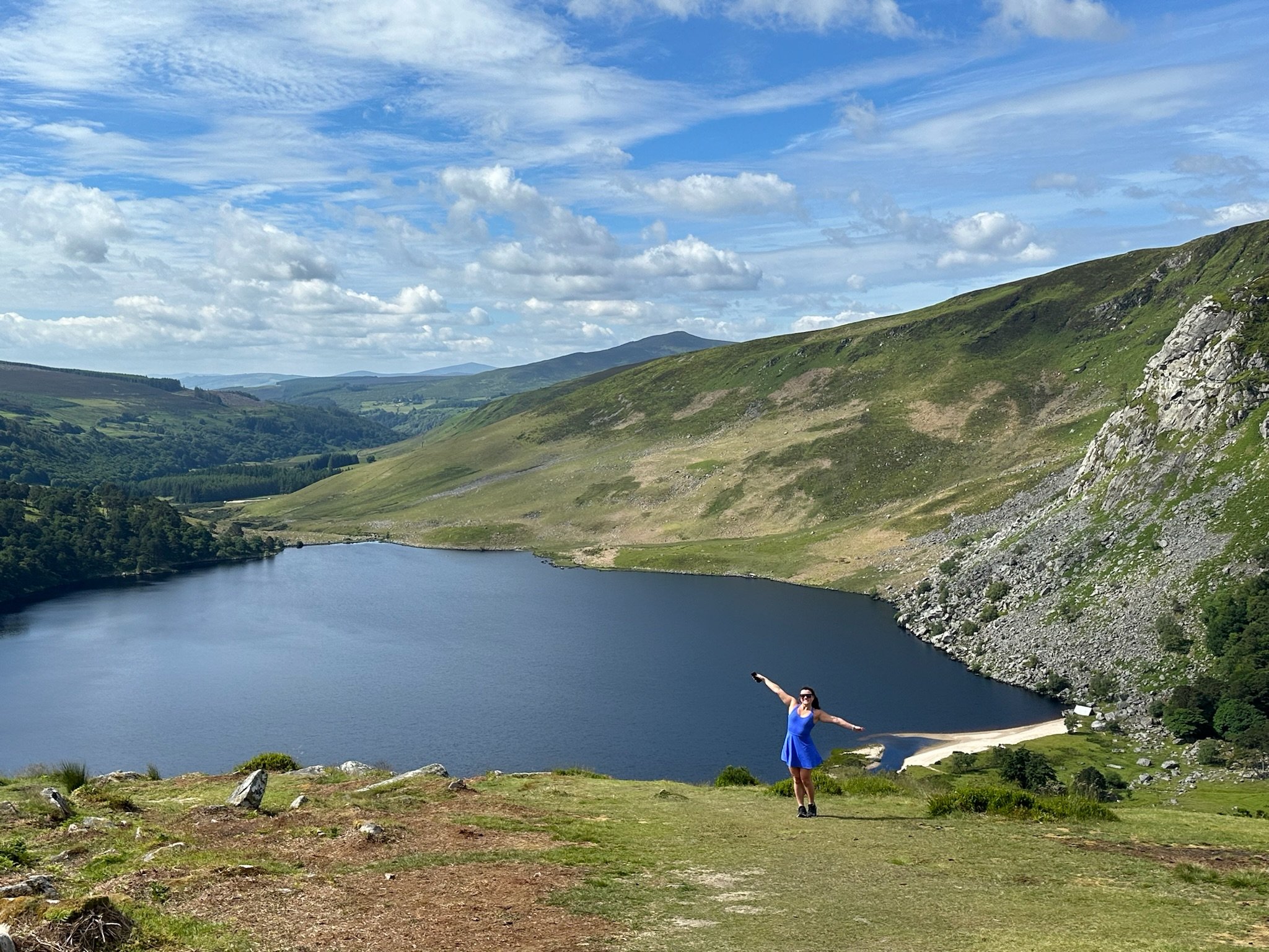 A woman in a blue dress standing by a lake with her arms outstretched, surrounded by green hills and mountains under a partly cloudy sky.
