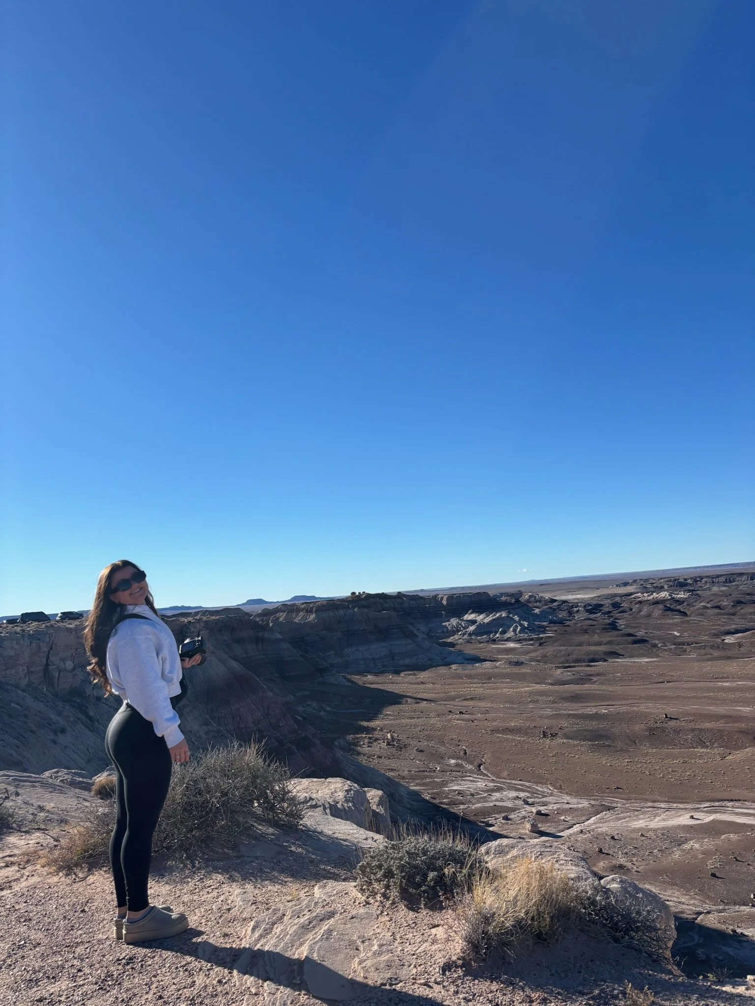 A woman with long hair wearing sunglasses, a white hoodie, black pants, and beige shoes standing in a desert landscape with rock formations under a clear blue sky.