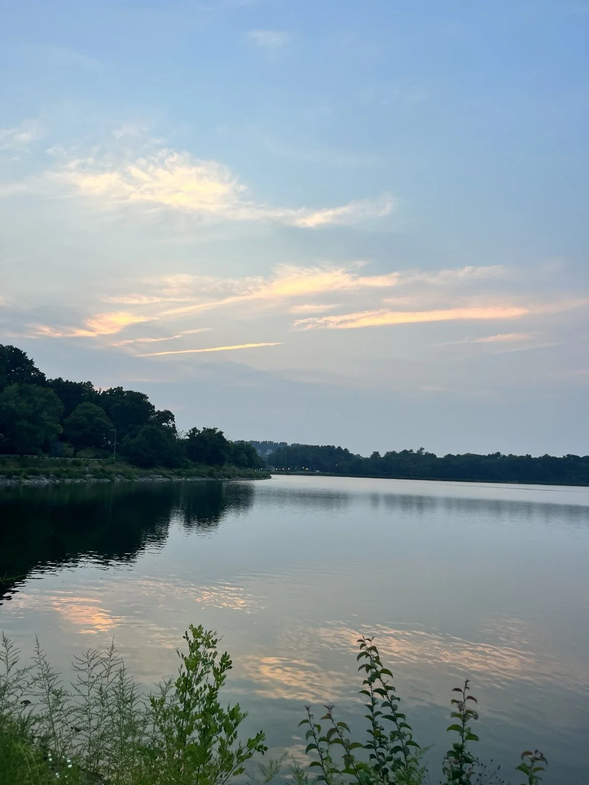 A calm river reflecting a partly cloudy sky at sunset or sunrise, with trees lining the riverbank.