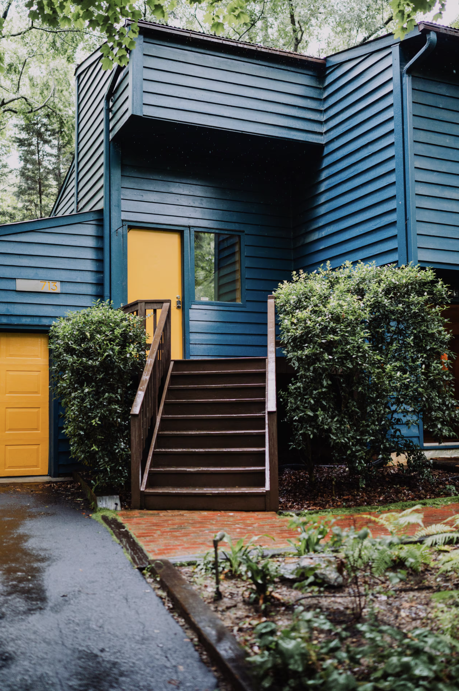 A blue wooden house with a yellow door, surrounded by green trees and bushes, with wooden steps leading up to the entrance, on a wet brick and asphalt pathway.