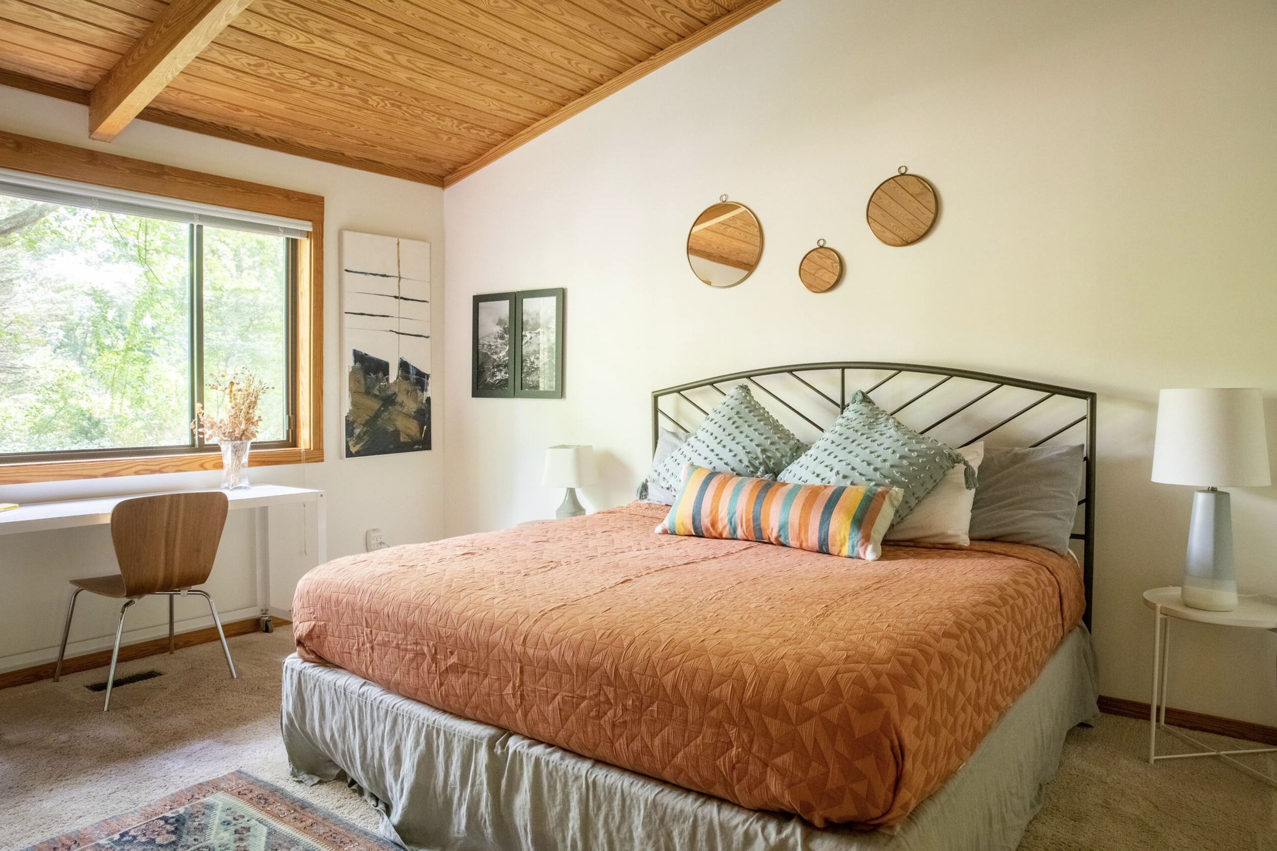 A cozy bedroom with a bed covered in orange quilt and colorful pillows, two nightstands with lamps, a window with green trees outside, decorative mirrors on the wall, and wooden ceiling beams.