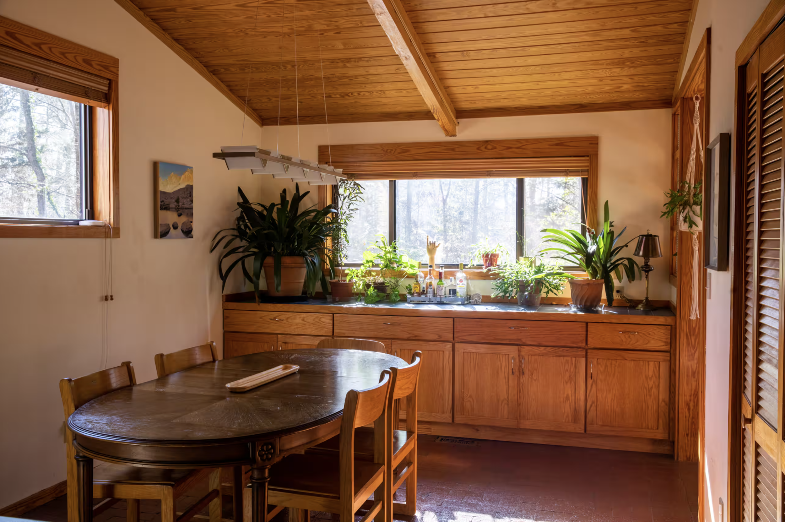 Wooden kitchen with large window, green plants, and a round dining table with chairs.
