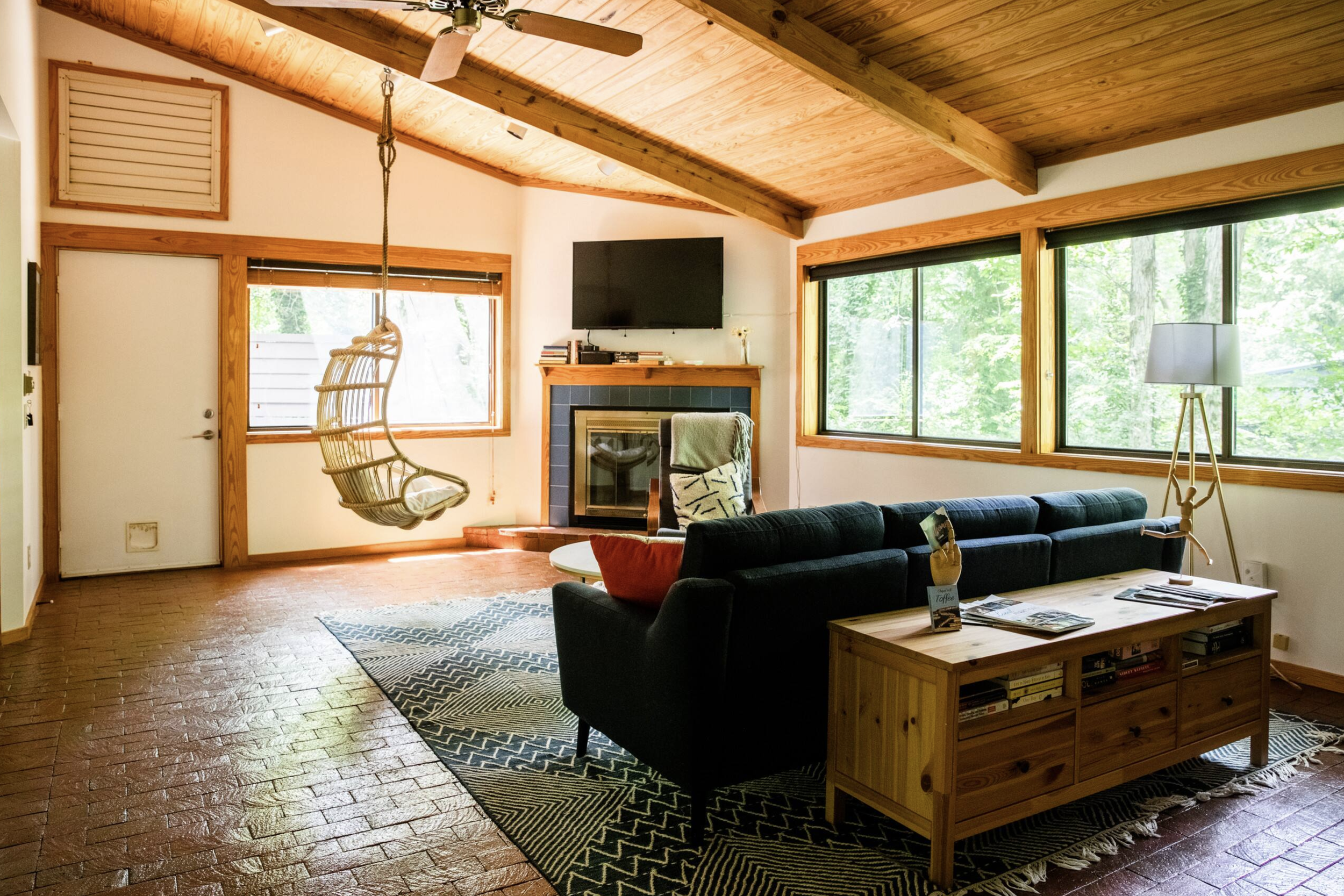 Living room with wooden ceiling and large windows showing green trees outside, a hanging chair, a flat-screen TV above a fireplace, and a sofa with a wooden coffee table and a bookshelf underneath.