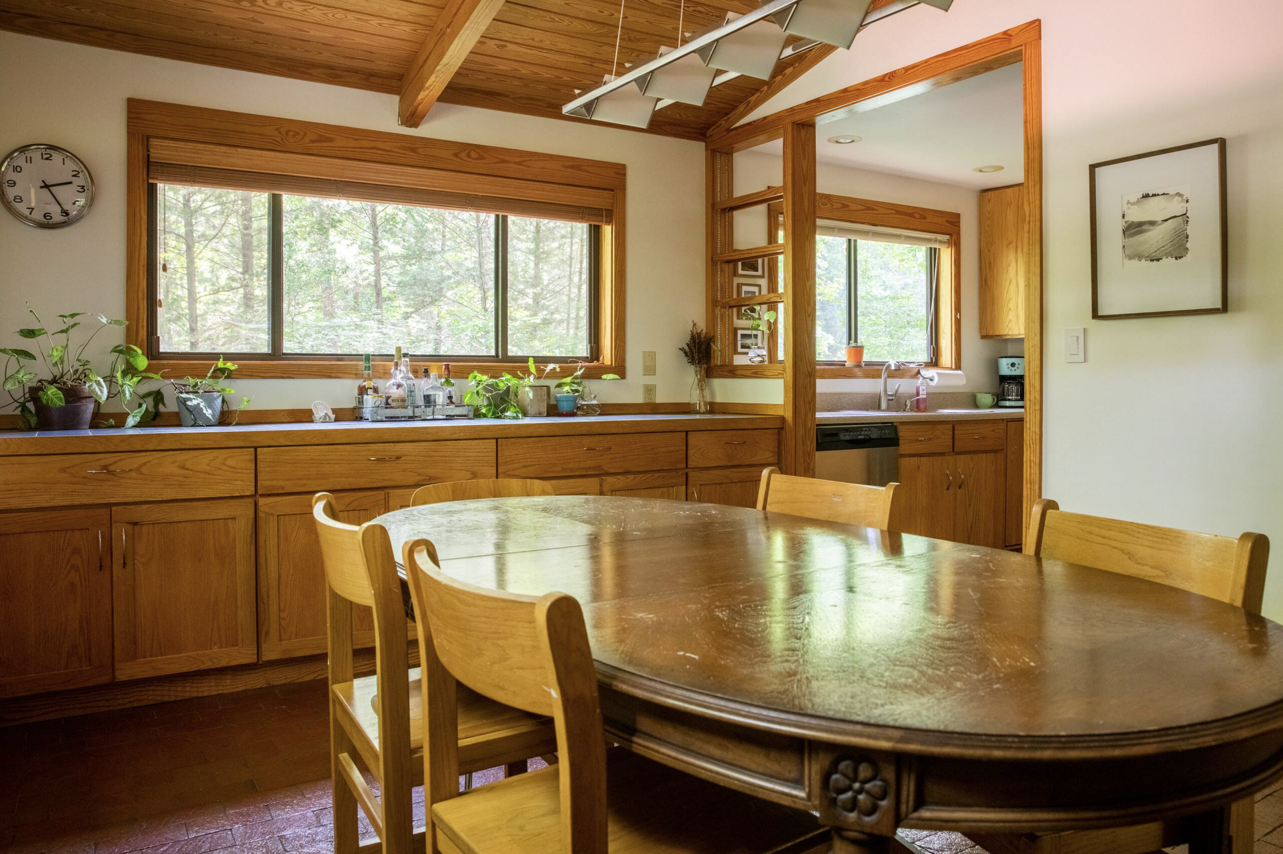 Wood-paneled kitchen with a large oval wooden dining table, several chairs, and windows showing a forest view. Potted plants and bottles on the counter, artwork on the wall, and natural light.