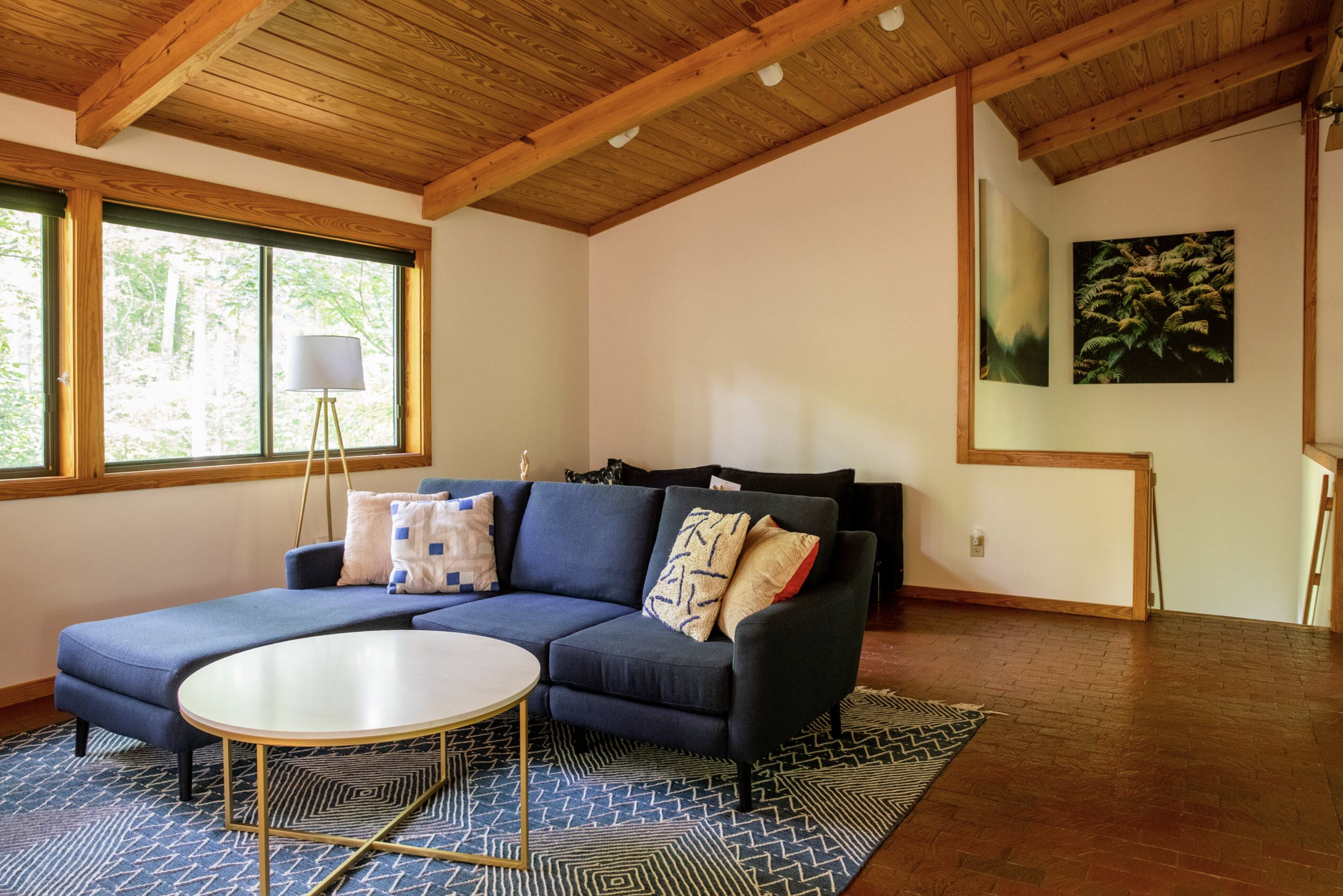 Living room with large window, dark blue sectional sofa with decorative pillows, round white coffee table, patterned rug, and wall art of greenery. Wooden ceiling with beams and trim.