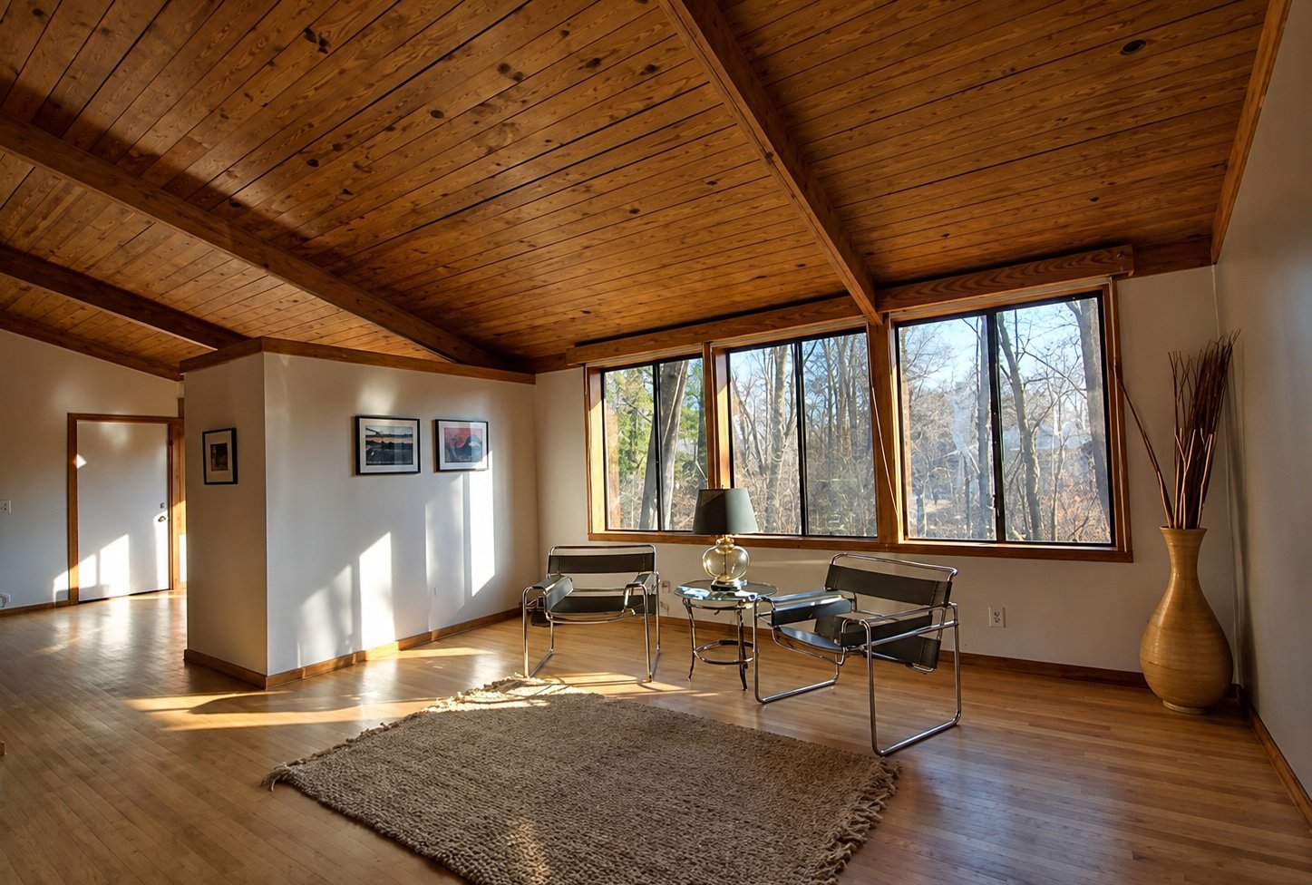 A living room with wood-paneled ceiling and large windows showing trees outside. There are two modern black and chrome chairs, a small glass table with a lamp, a large beige rug, and a tall decorative vase with sticks.