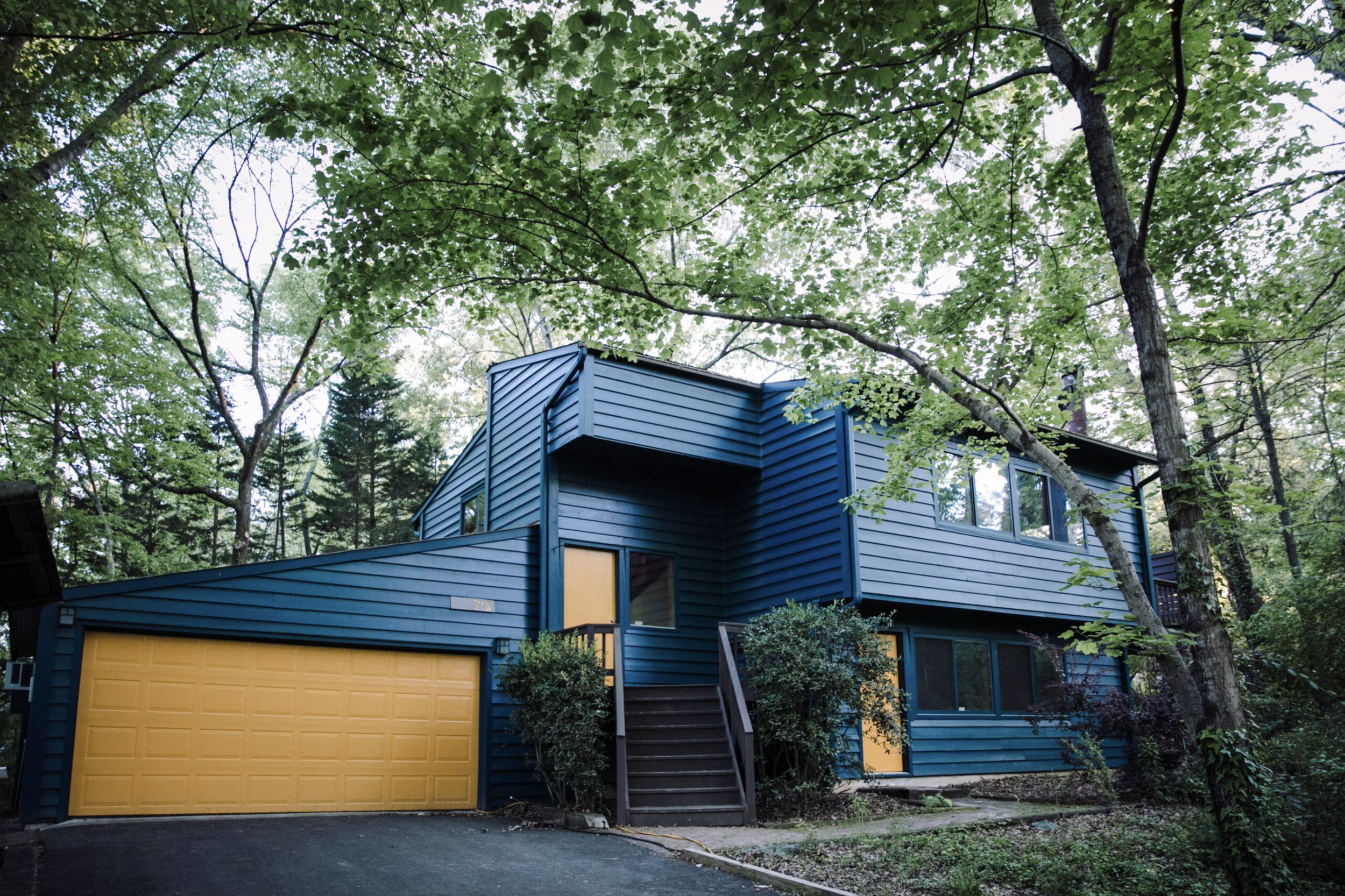 A modern blue house with a yellow garage door is surrounded by tall trees with green leaves, in a wooded area.