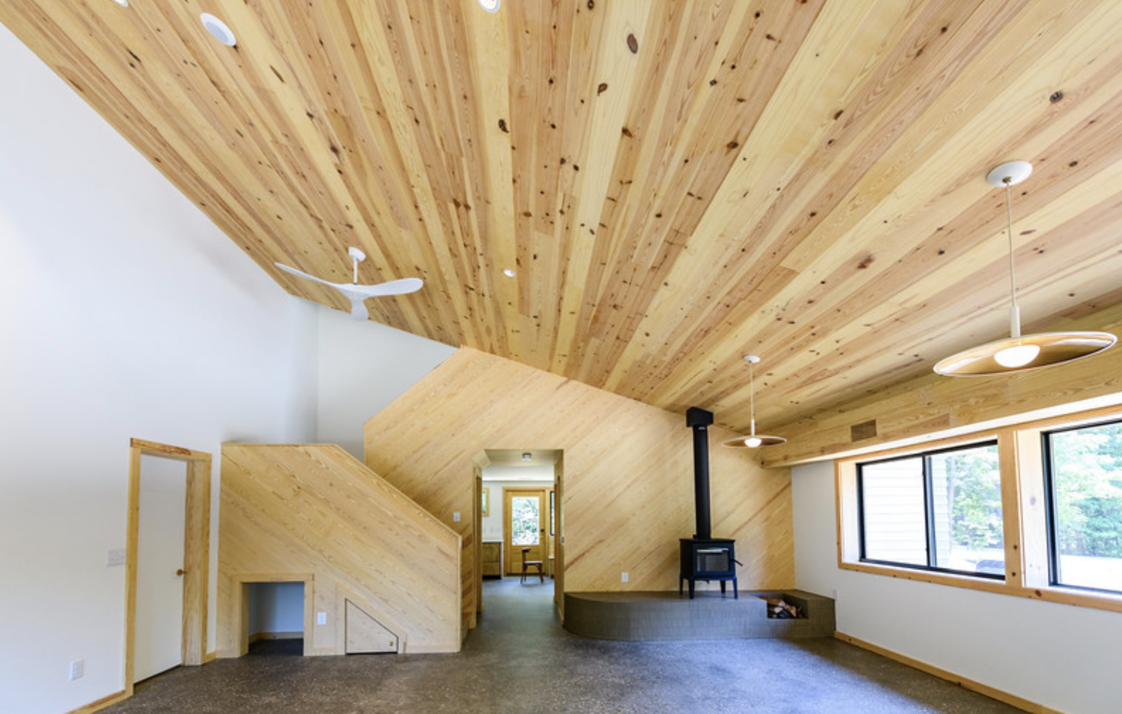 Interior view of a modern home with a curved wooden ceiling, a wood stove, hanging pendant lights, large windows, and light-colored walls.