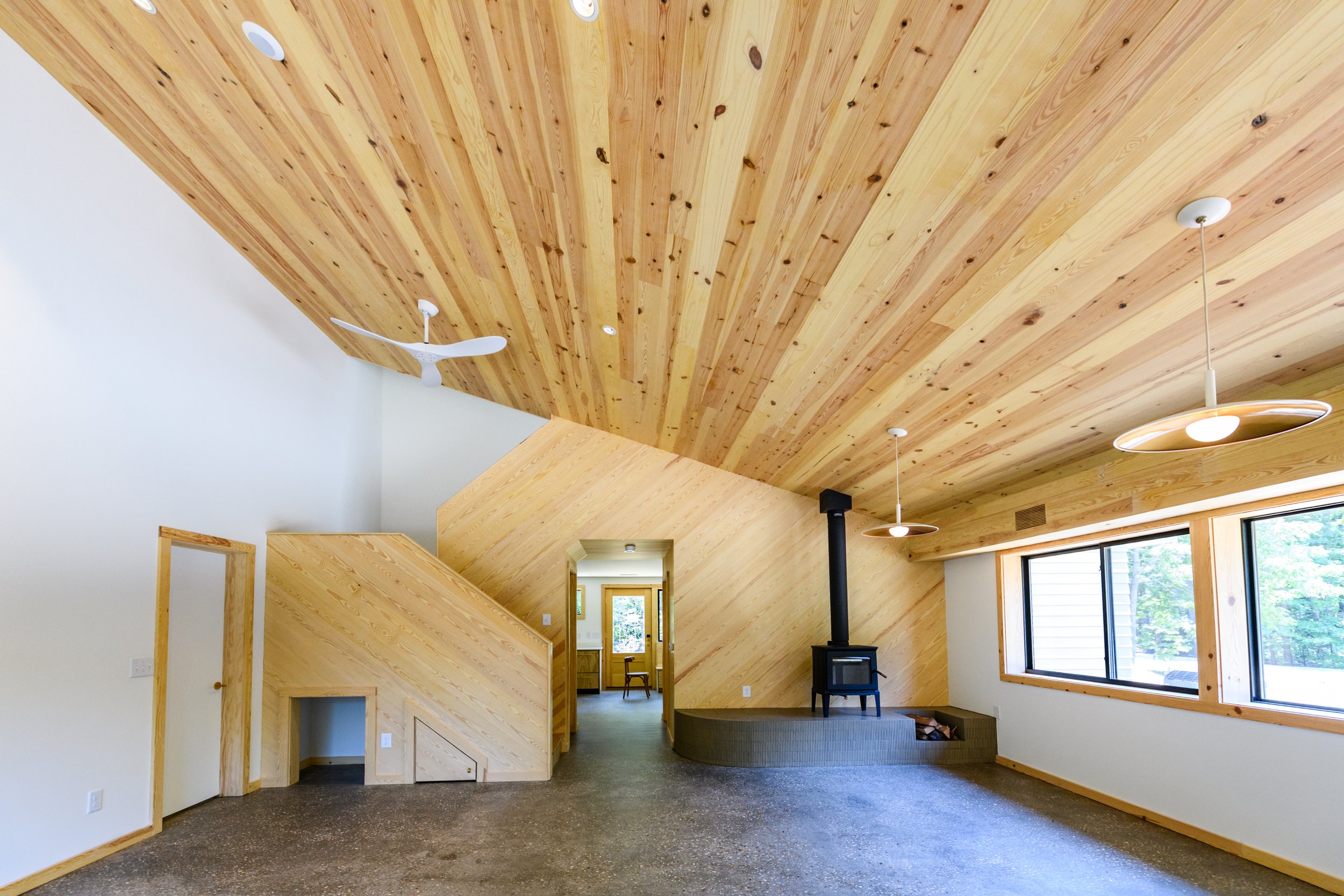 Interior living space with wood vaulted ceiling, large windows, a black wood stove, ceiling fans, and contemporary lighting fixtures.