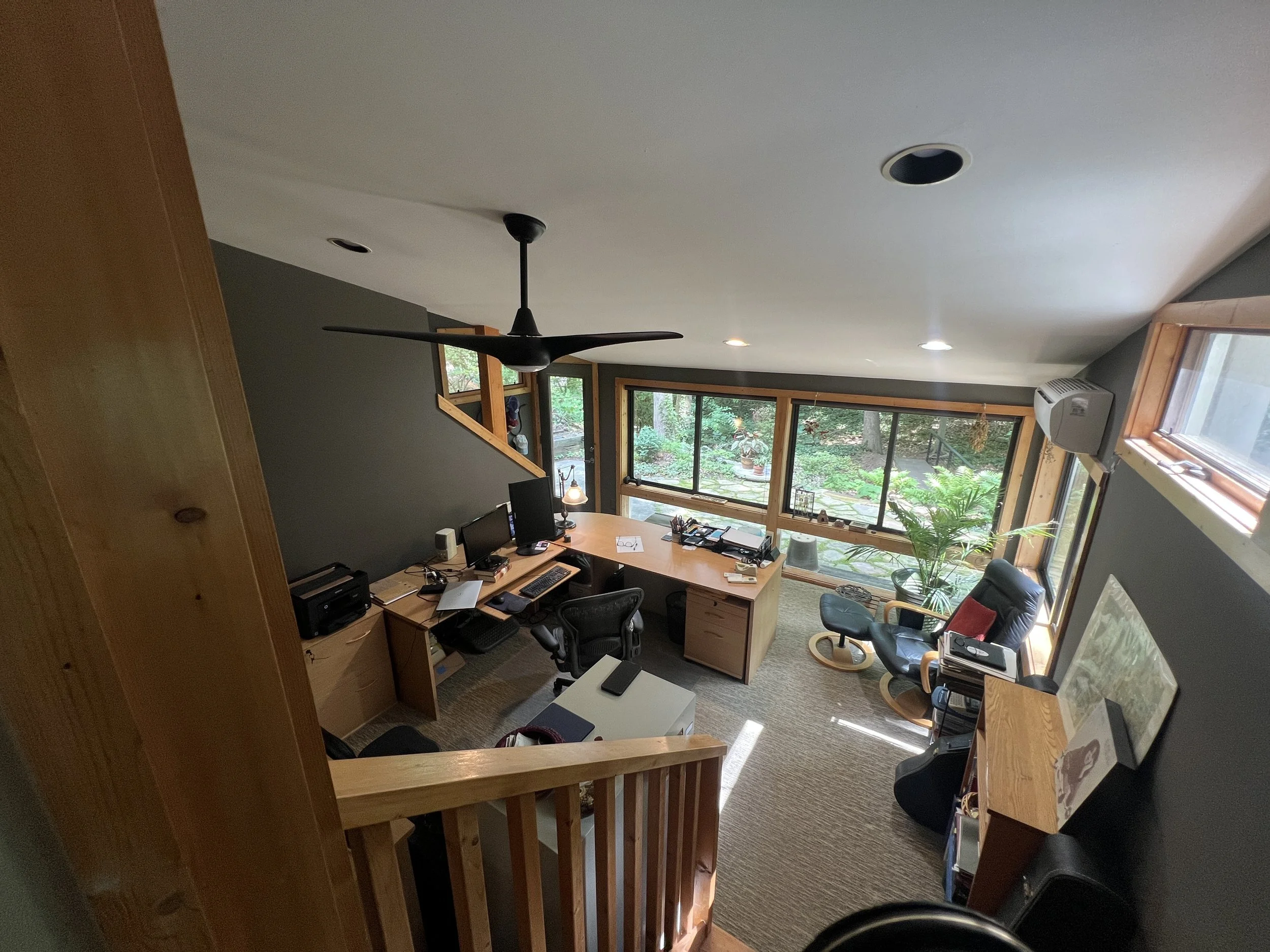 Interior view of a home office with a large window overlooking a wooded outdoor area. The ceiling has two recessed lights and a ceiling fan. The room contains a wooden desk with multiple monitors, office supplies, and a black office chair. There are 