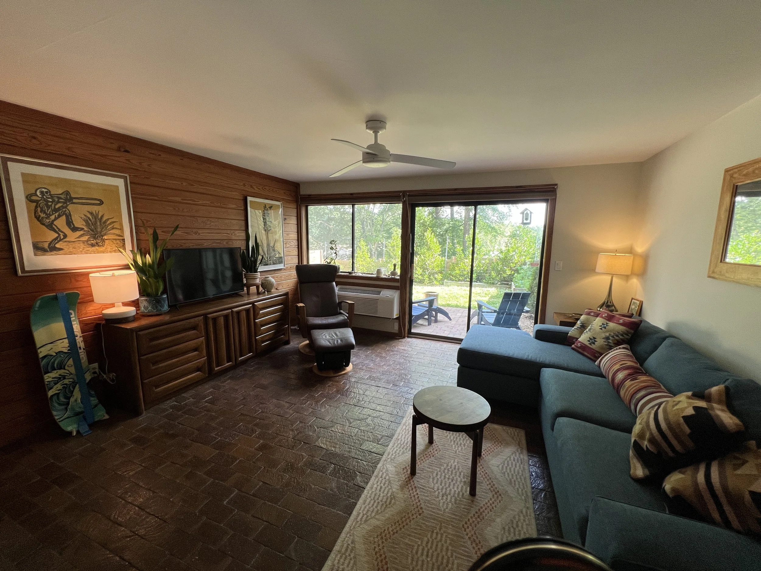 Living room with brick flooring, wooden-paneled wall, a window with a sliding door leading outside, a ceiling fan, a blue sofa with pillows, a small round table, a tall floor lamp, a TV on a wooden stand, indoor plants, and artwork on the wall.