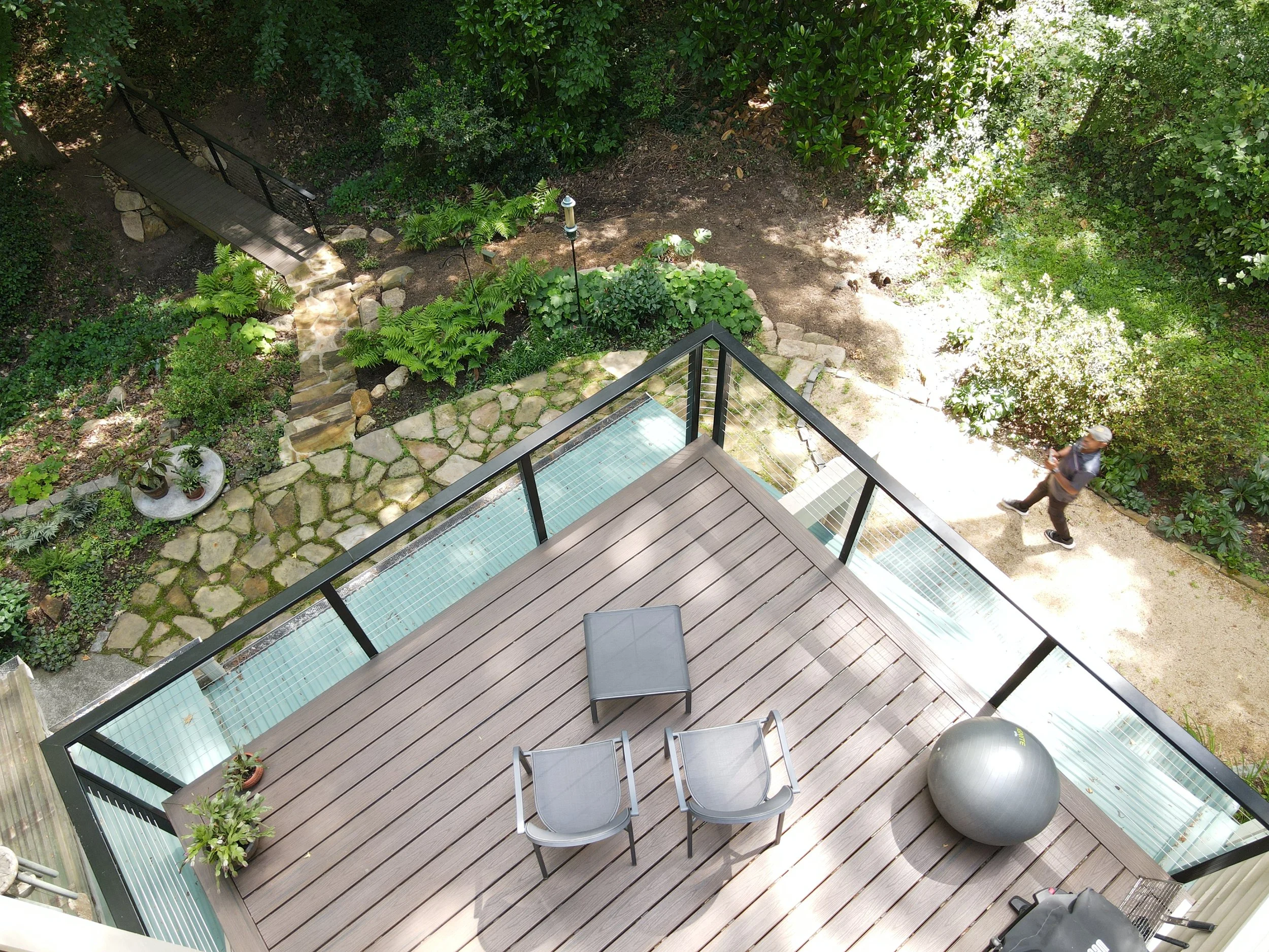 A wooden balcony with a black chair and a large exercise ball, overlooking a garden with a stone pathway, potted plants, and lush green trees and bushes.