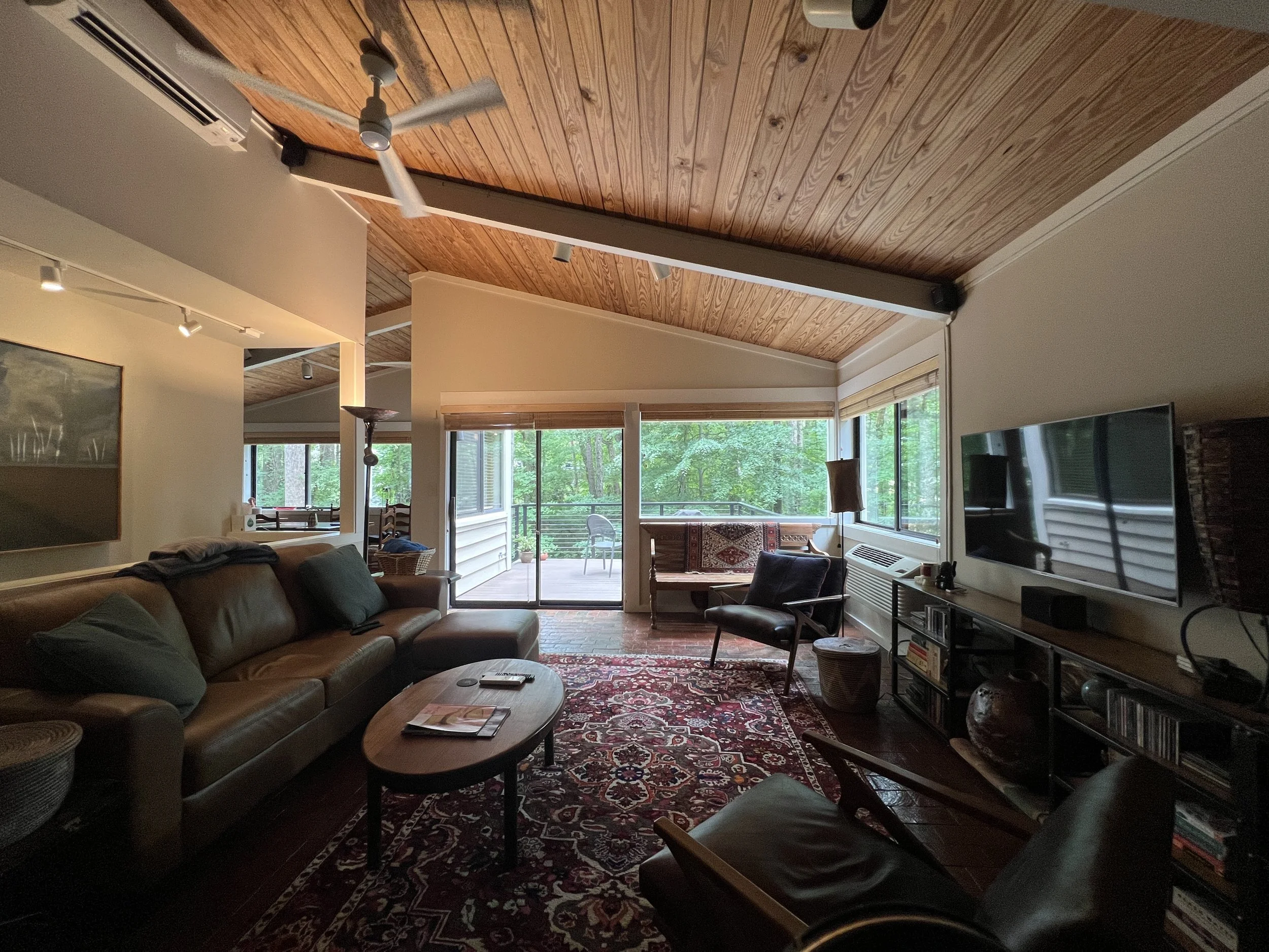 Living room with brown leather sofa, wooden ceiling, large windows showing green trees outside, rug, various chairs, television, and home decor.