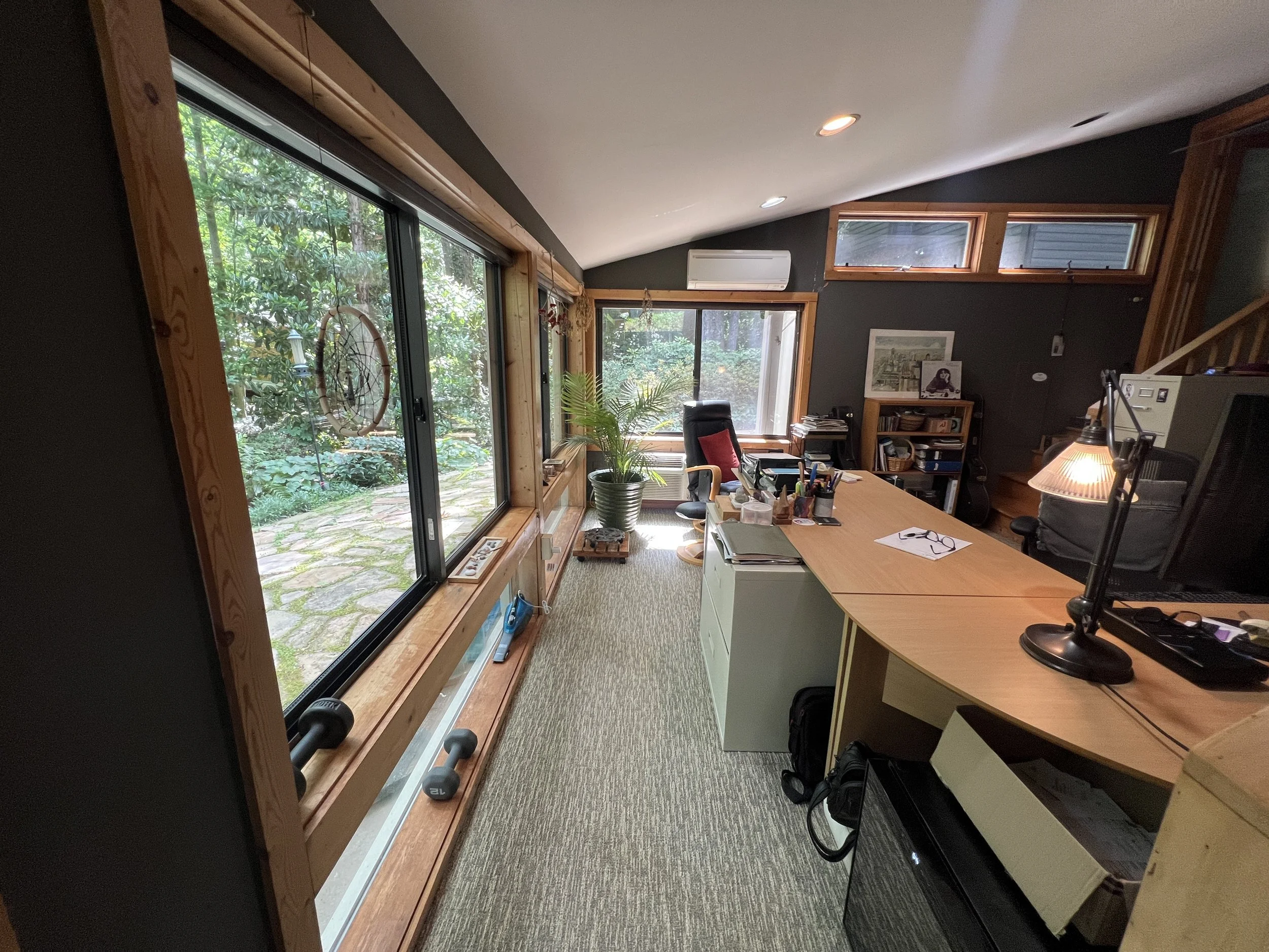 Home office with large windows overlooking a green garden, a wooden desk with office supplies, a black office chair, and framed artwork on dark walls.