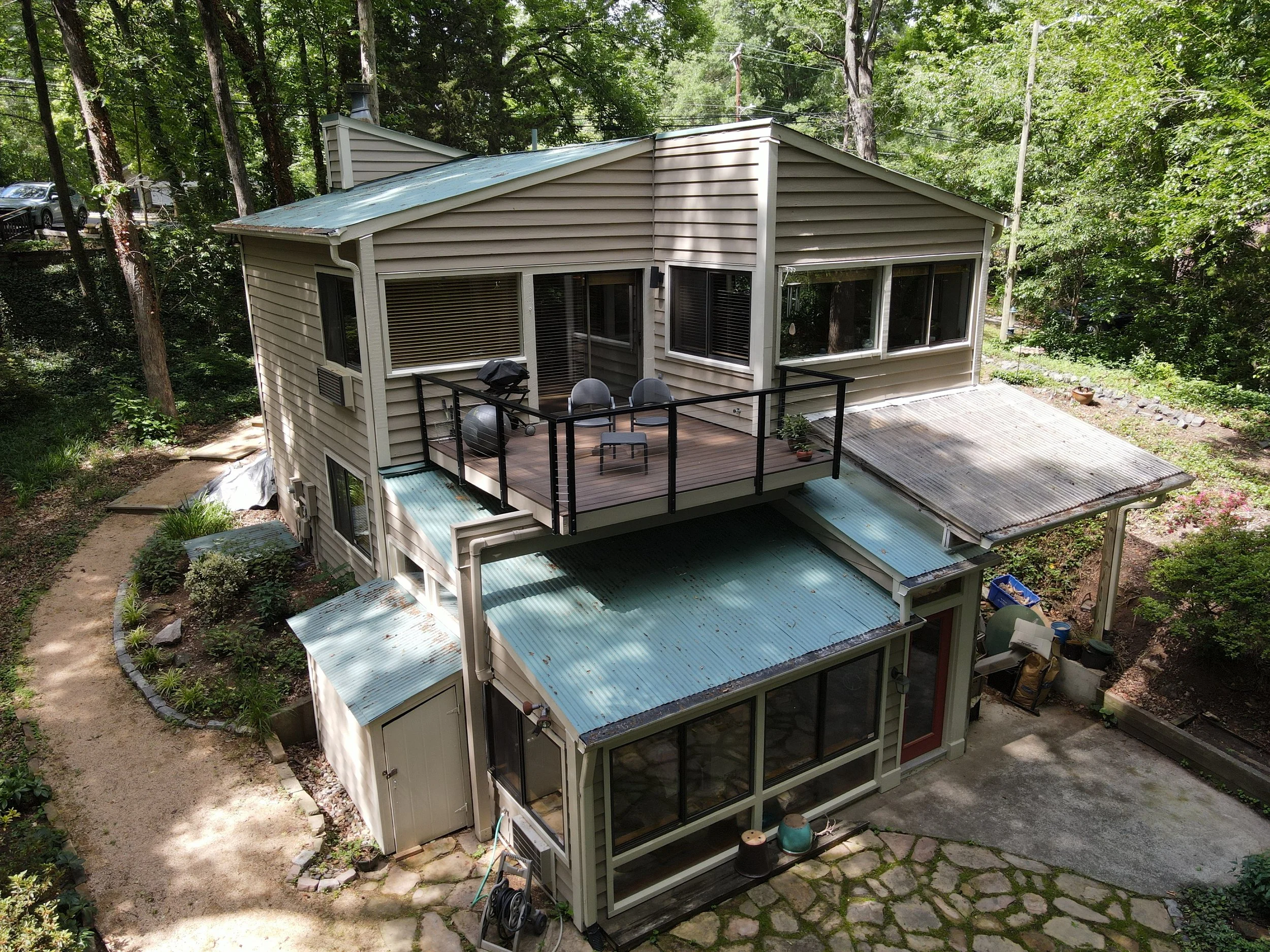 A multi-story house in a wooded area featuring large glass windows, a balcony with chairs, and a metal roof, with trees and a patio surrounding it.