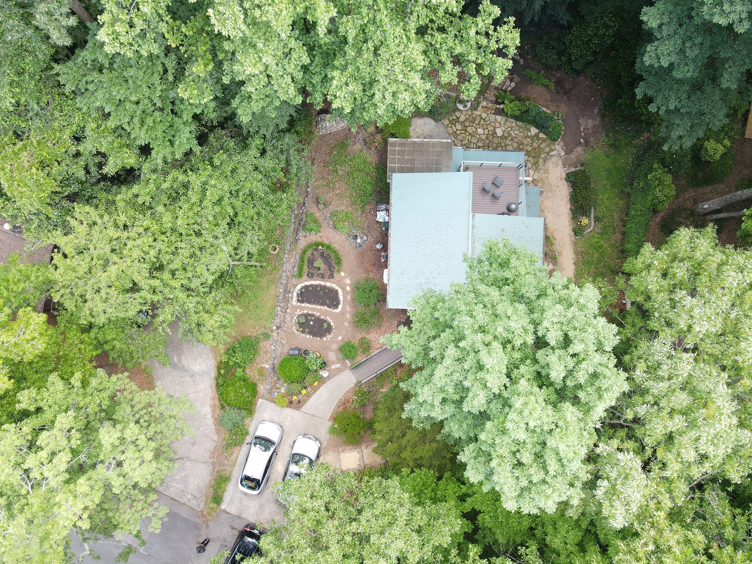 An aerial view of a backyard with green trees, a stone pathway, patches of soil, and parked cars.
