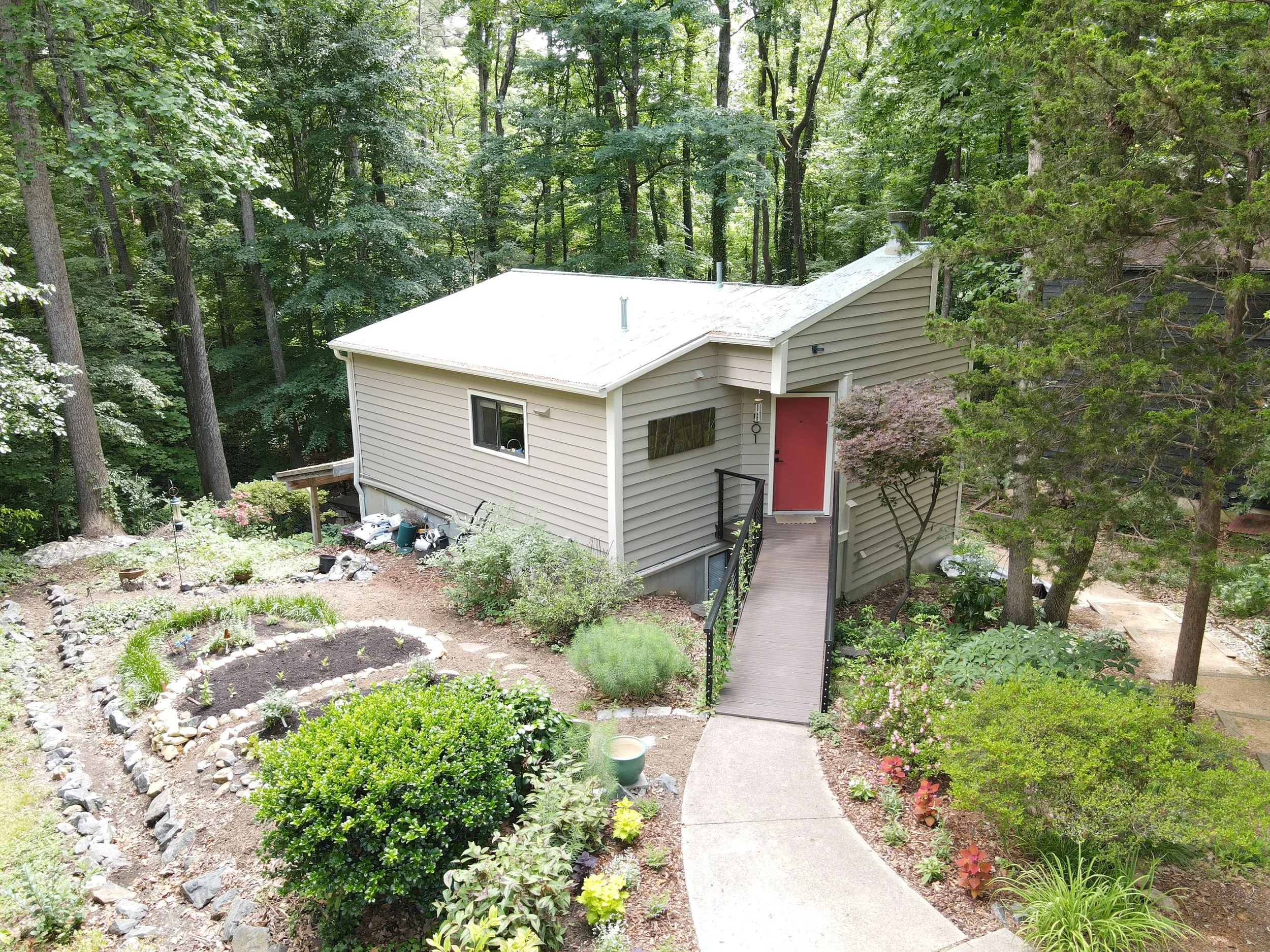 A view of a beige house with a red front door and a ramp, surrounded by a lush green garden with trees, plants, and a pathway, with a forested area in the background.