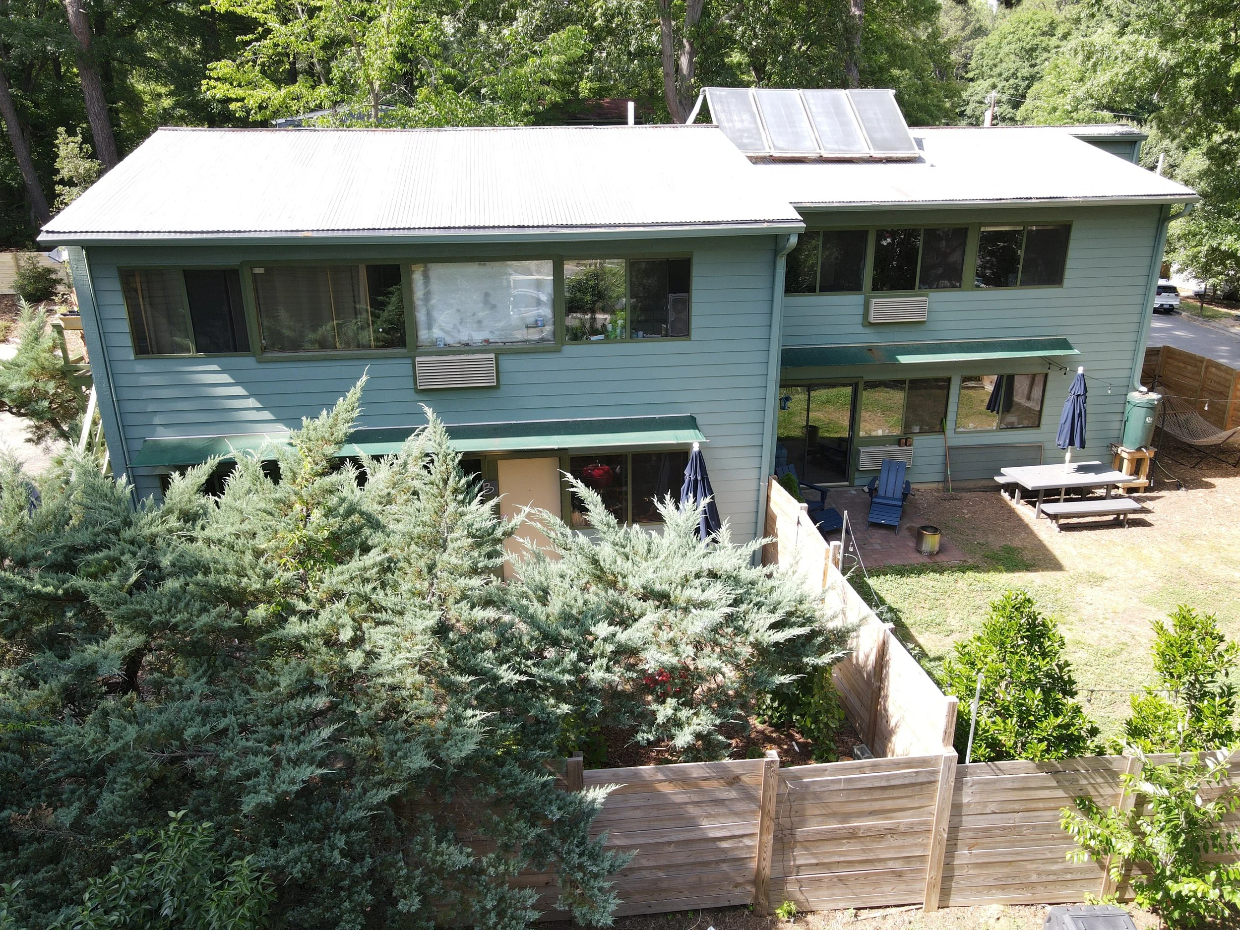A two-story house with light blue siding, green window frames, and a white metal roof. Solar panels are installed on part of the roof. The backyard has a patio with outdoor furniture, umbrellas, and a wooden privacy fence. The area is surrounded by t
