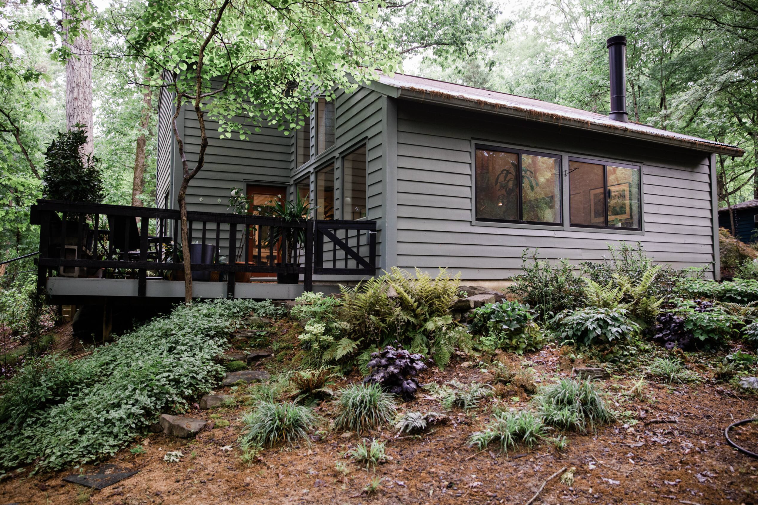A house surrounded by lush green trees and plants, featuring large windows, a deck with potted plants, and a forest setting.