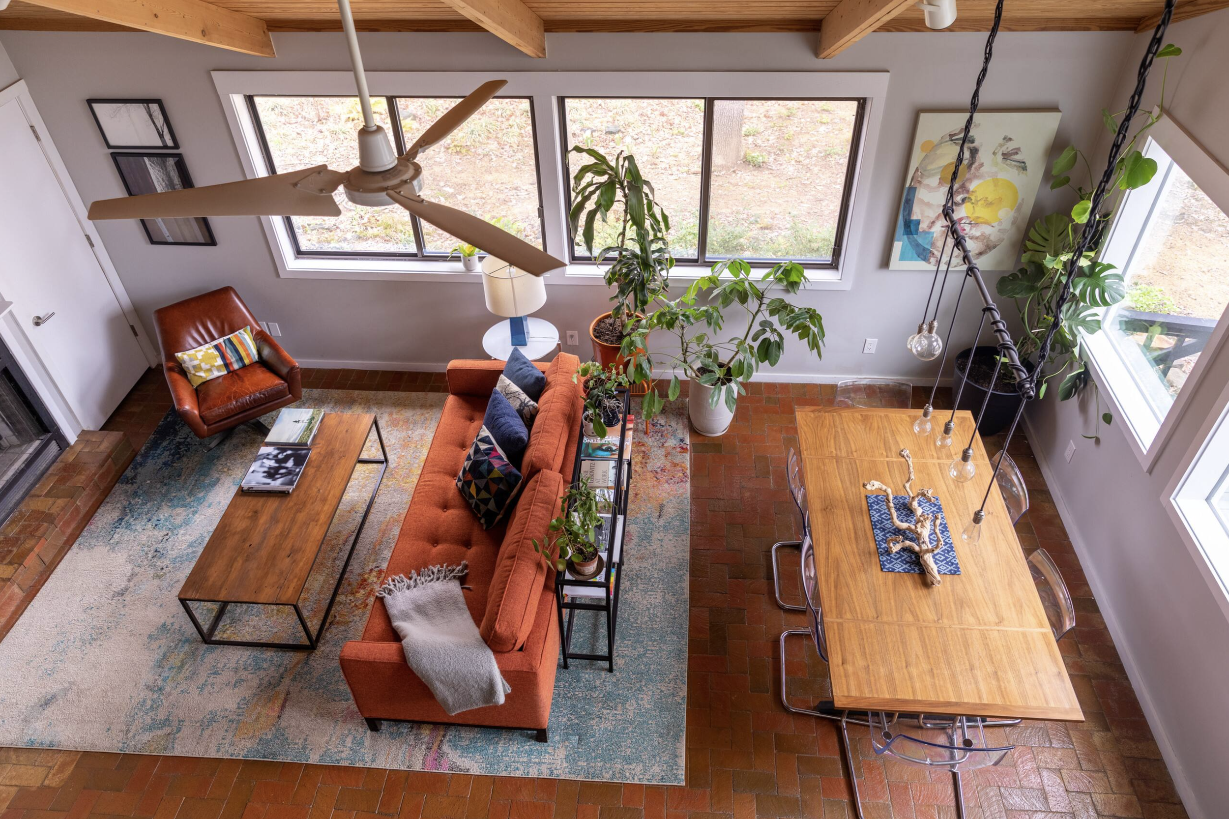 A living room with large windows, a ceiling fan, a red leather armchair, a sofa with colorful pillows, a coffee table, a rug, and indoor plants near the windows.