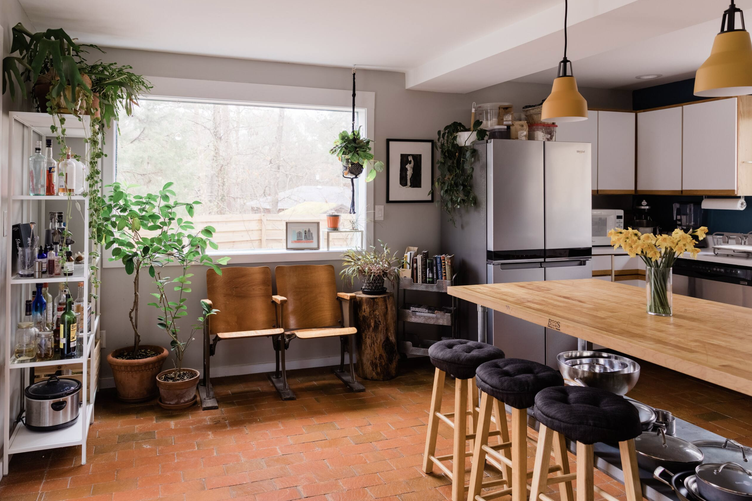 A cozy kitchen and dining area with a large window, potted plants, a wooden table with black cushioned stools, a white shelving unit with bottles and jars, and a refrigerator, with pendant lights hanging from the ceiling.