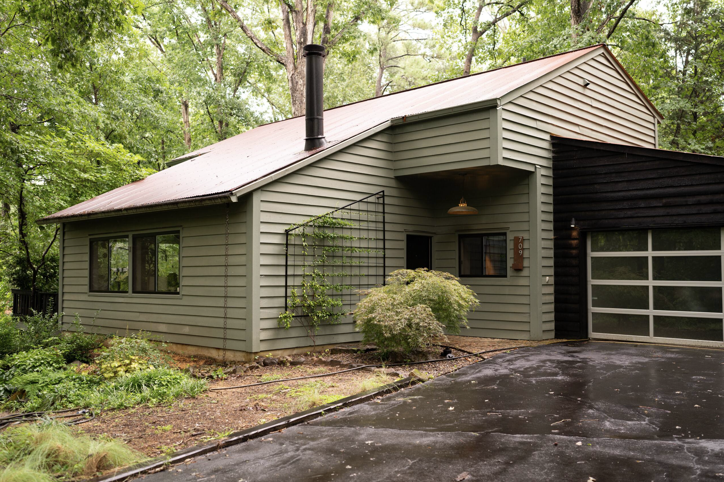 A modern house with beige and black exterior siding, surrounded by green trees and plants, with a driveway in the foreground.
