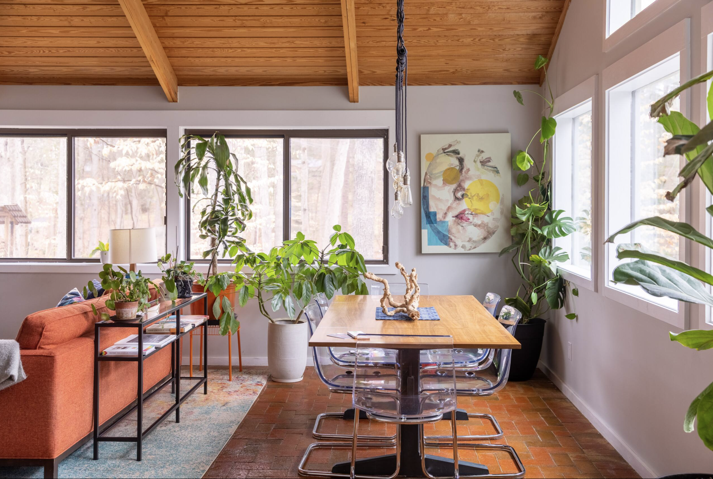 Bright living room with large windows, wooden ceiling, artwork on wall, numerous green houseplants, a wooden dining table with clear chairs, and a black metal side table with books, a lamp, and plants.