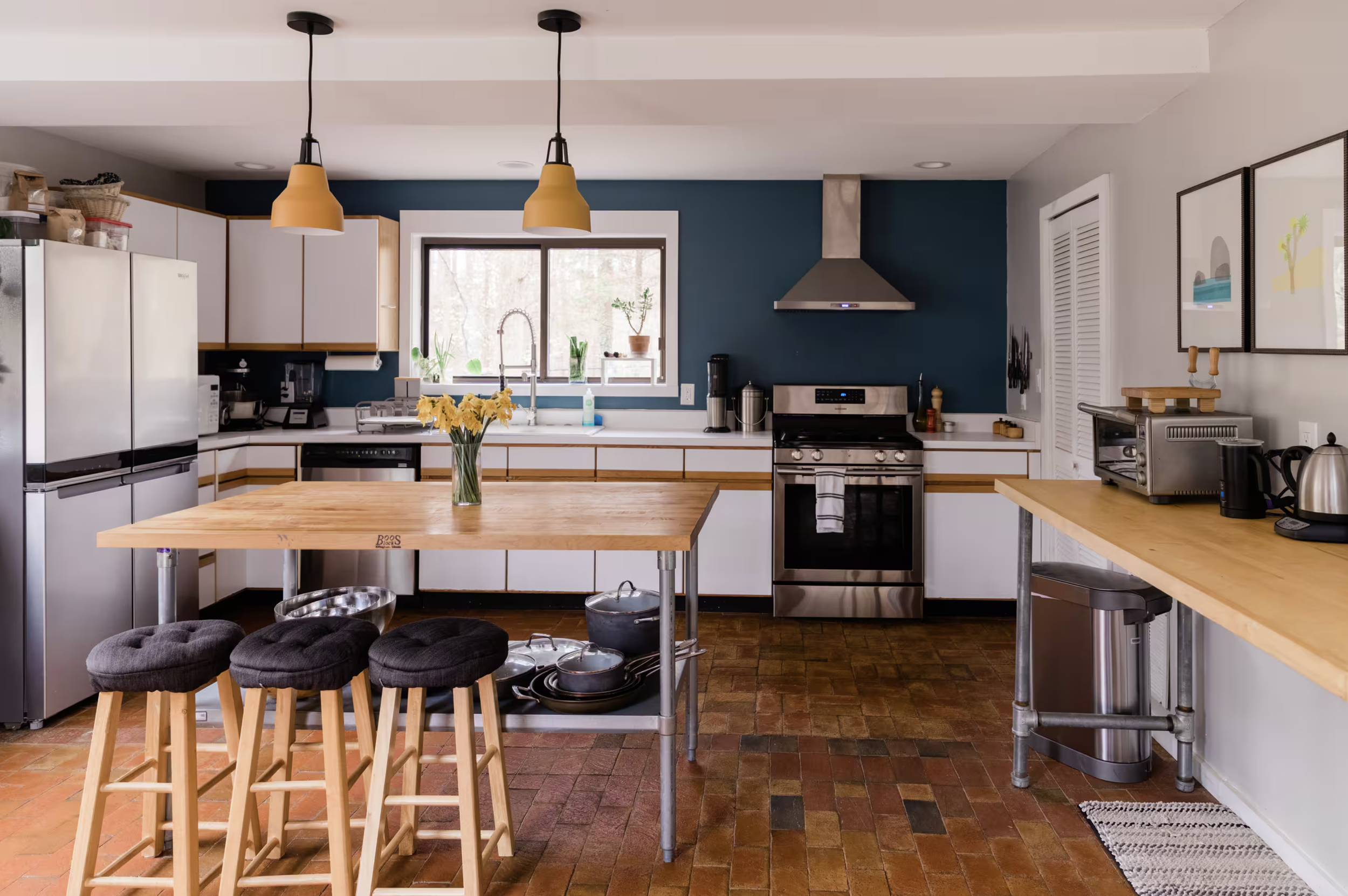 A modern kitchen with white cabinets and a dark blue accent wall. There are two yellow pendant lights hanging over a wooden island with three black stools. The kitchen features stainless steel appliances, including a refrigerator and oven. The counte